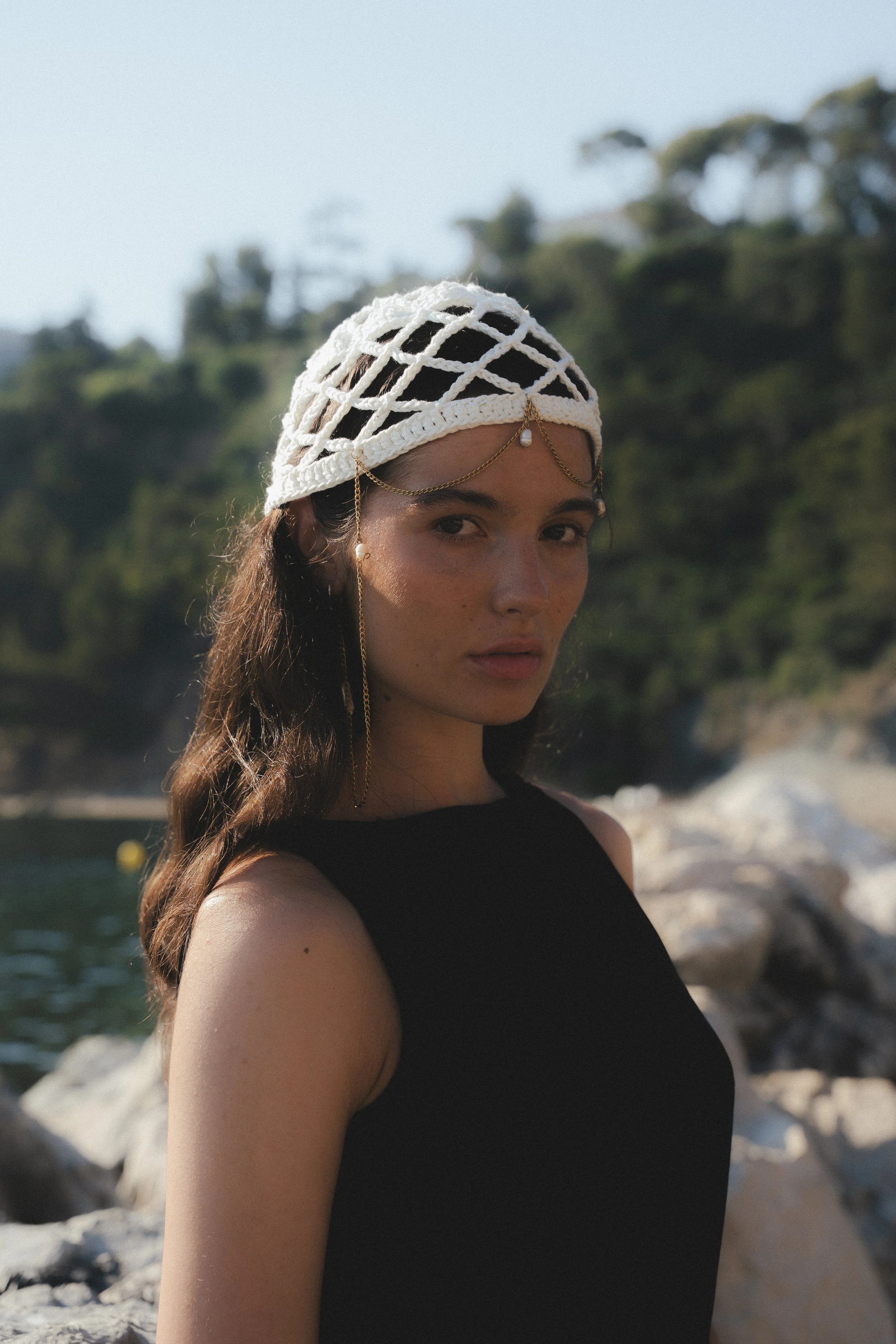 A young woman with long brown hair wearing a black sleeveless top and a white knitted headpiece with gold chains and pearl embellishments, standing near a rocky shoreline with trees in the background under a clear sky.