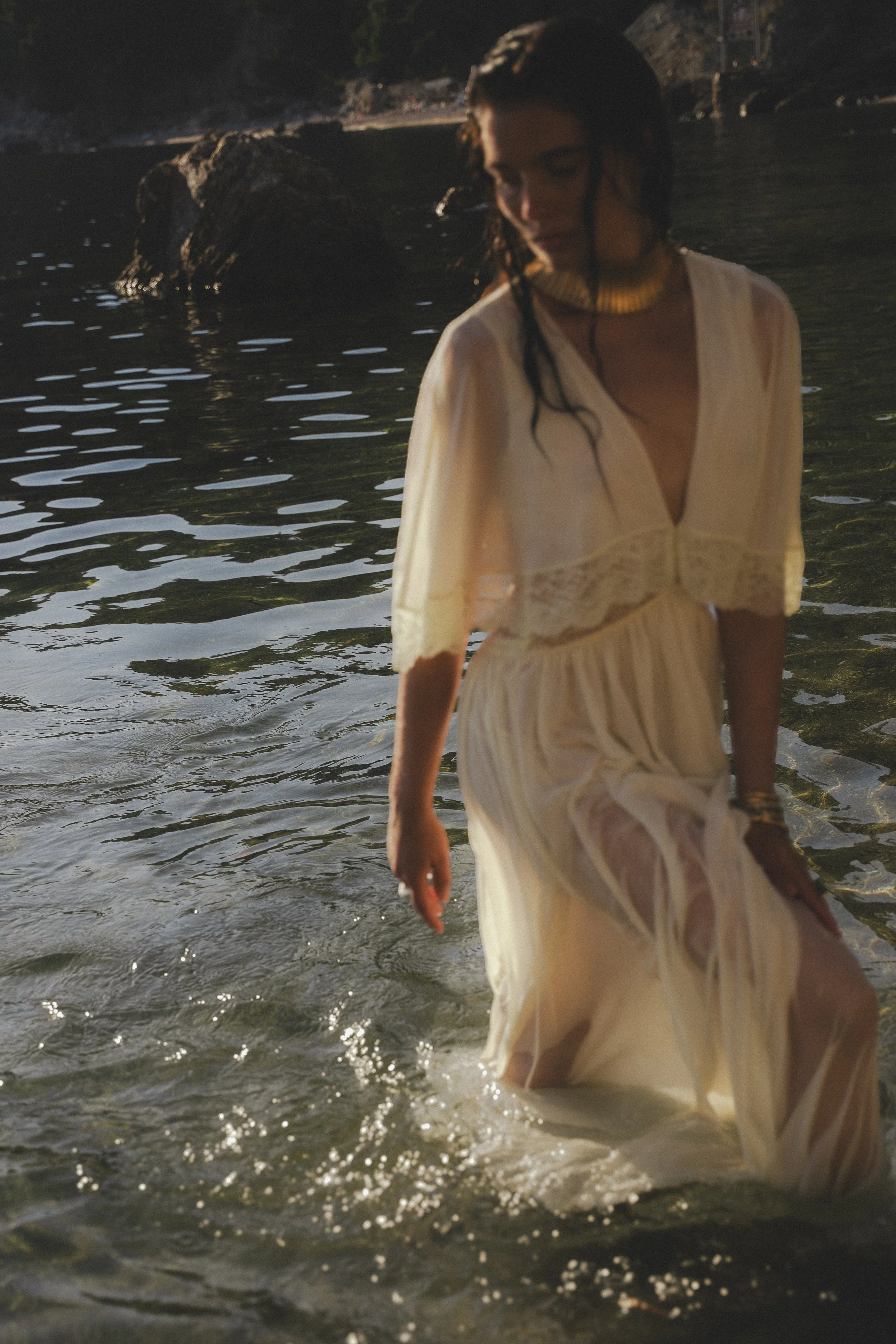 A woman in a light-colored dress walking in shallow water at the beach during sunset.