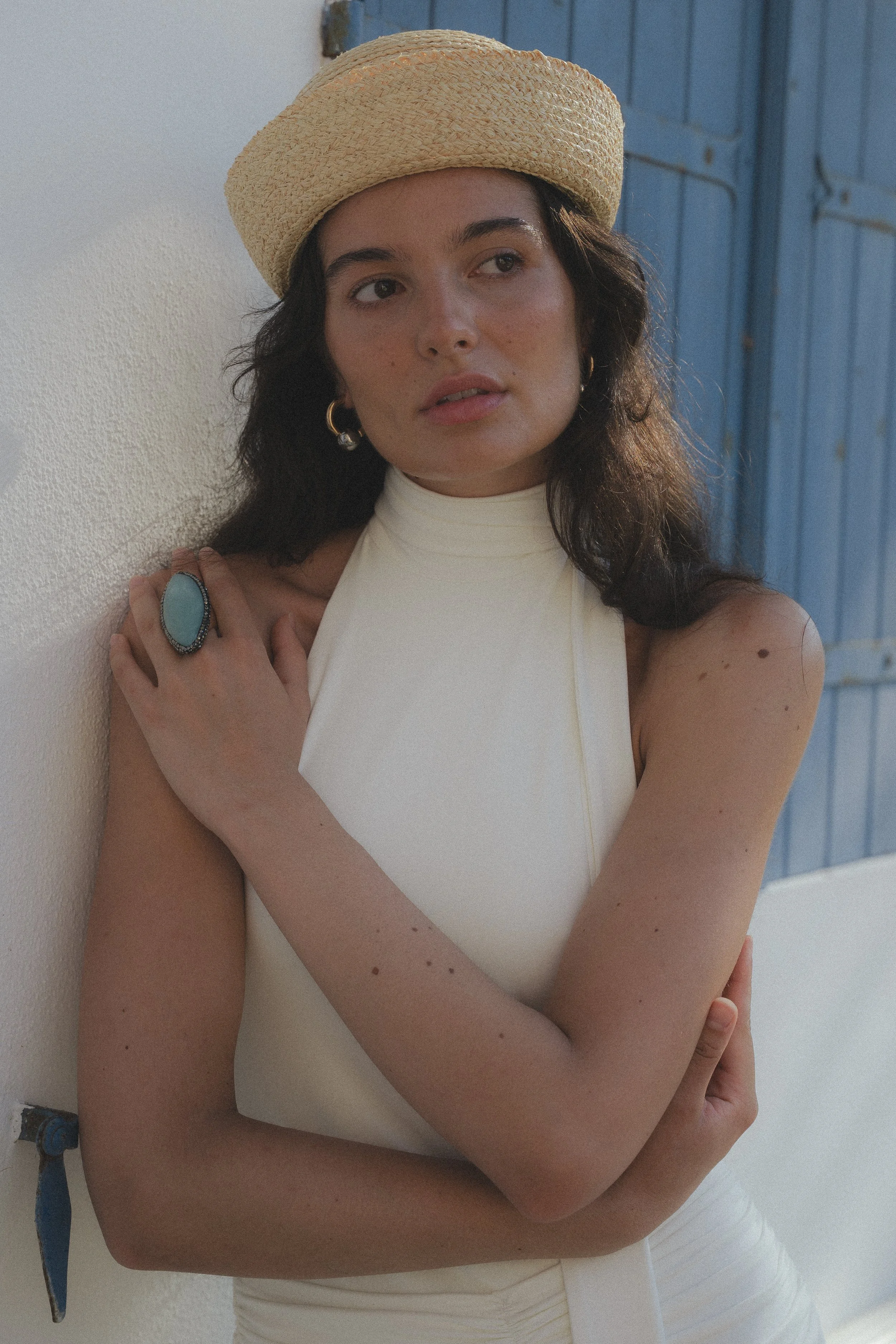 A young woman wearing a straw hat, gold hoop earrings, a large turquoise ring, and a sleeveless white top, standing against a blue wooden door and white wall, with her arms crossed.