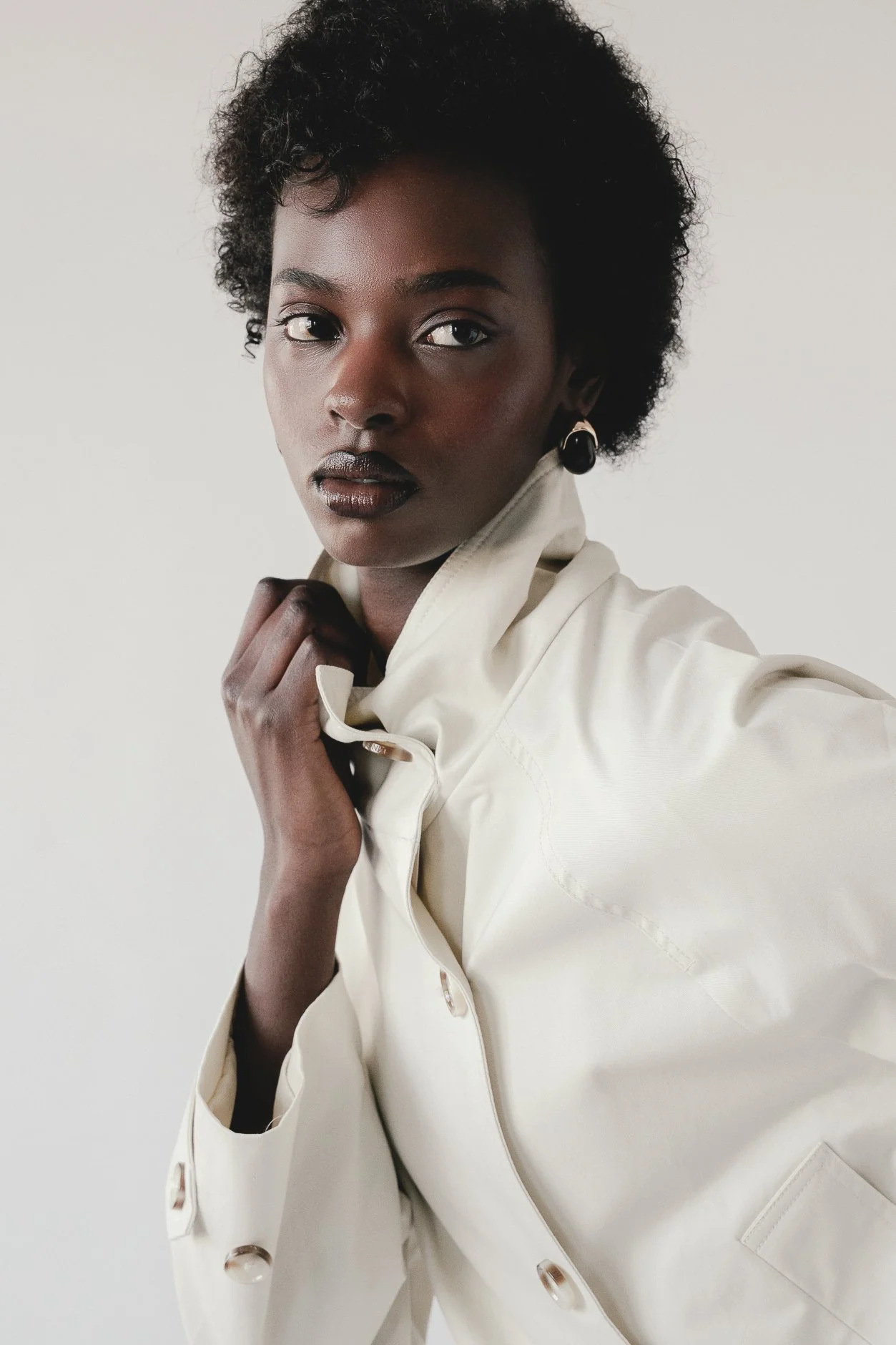 A woman with dark skin, short curly hair, wearing a cream-colored jacket and black earrings, looking directly at the camera with a neutral expression, against a plain white background.