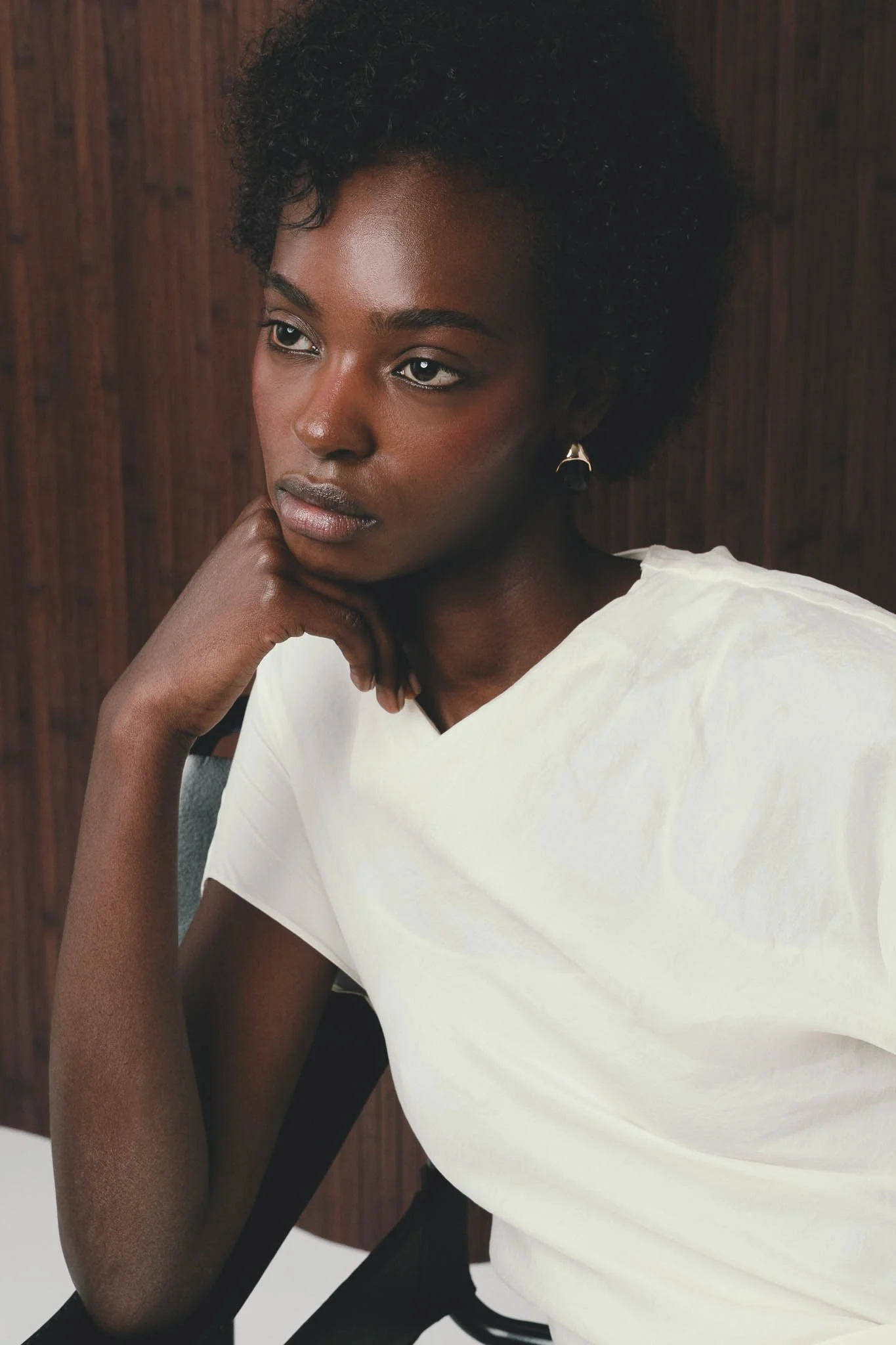 A woman with short, curly black hair and dark skin, resting her face on her hand, wearing a white shirt, and sitting on a black chair in front of a wooden background.