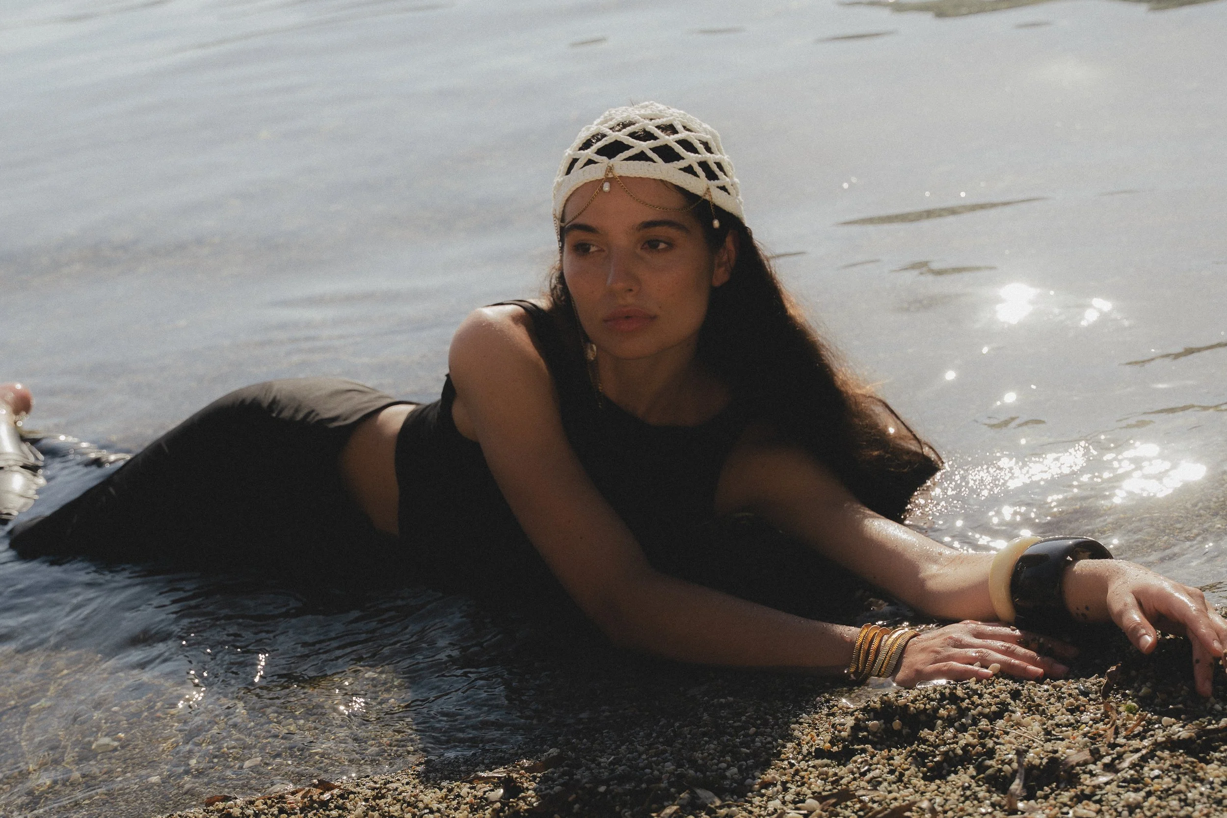 Woman lying on her stomach on the rocky shore, stretching with her arms extended forward, near the water's edge, wearing a woven hat and multiple bracelets.