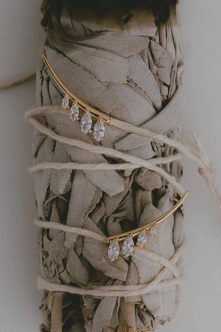 Gold earrings with five teardrop-shaped clear gemstones, wrapped around a bundle of dried gray leaves and twine.