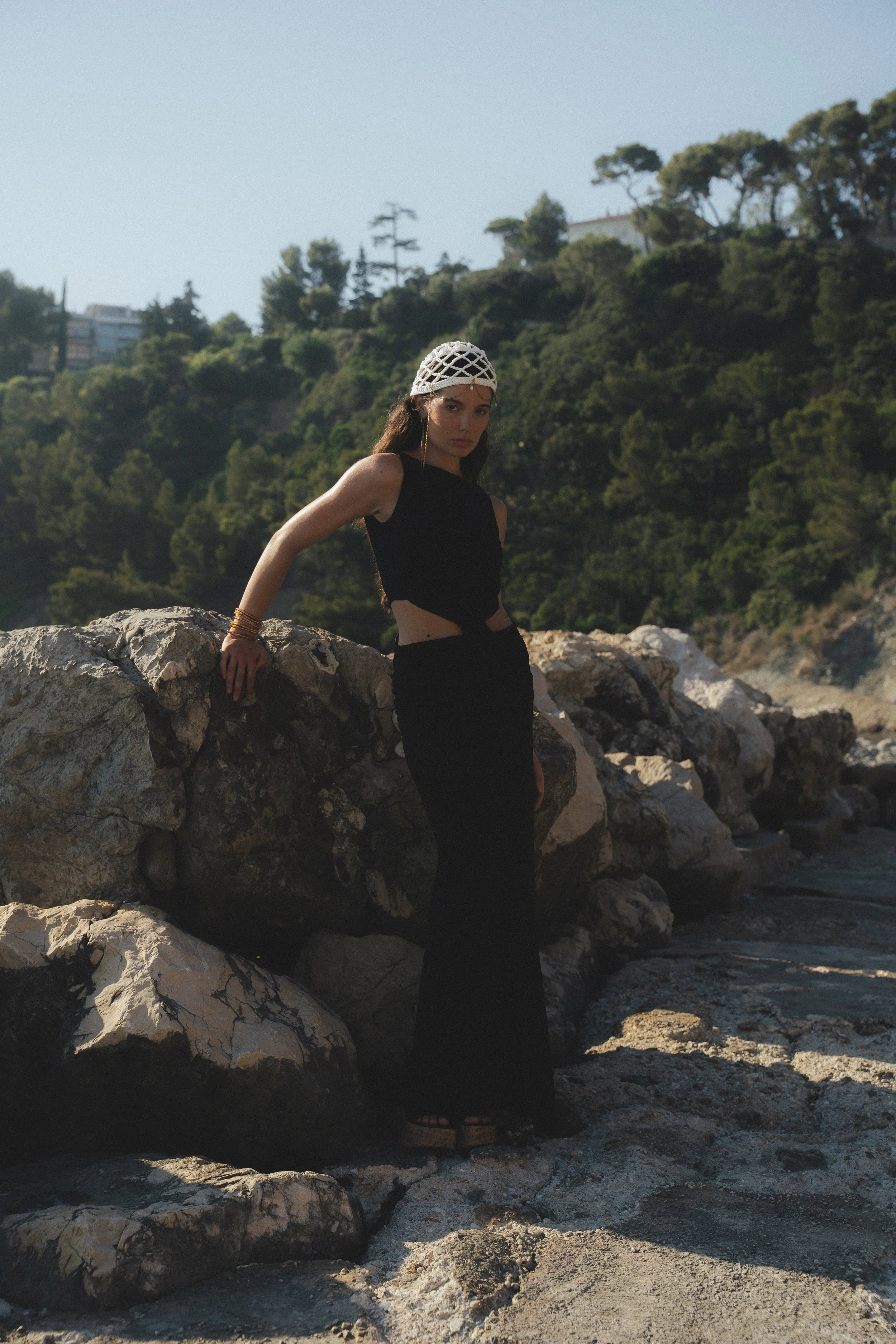 A woman in a black dress and a white head covering standing among large rocks near a beach, with greenery and trees in the background.