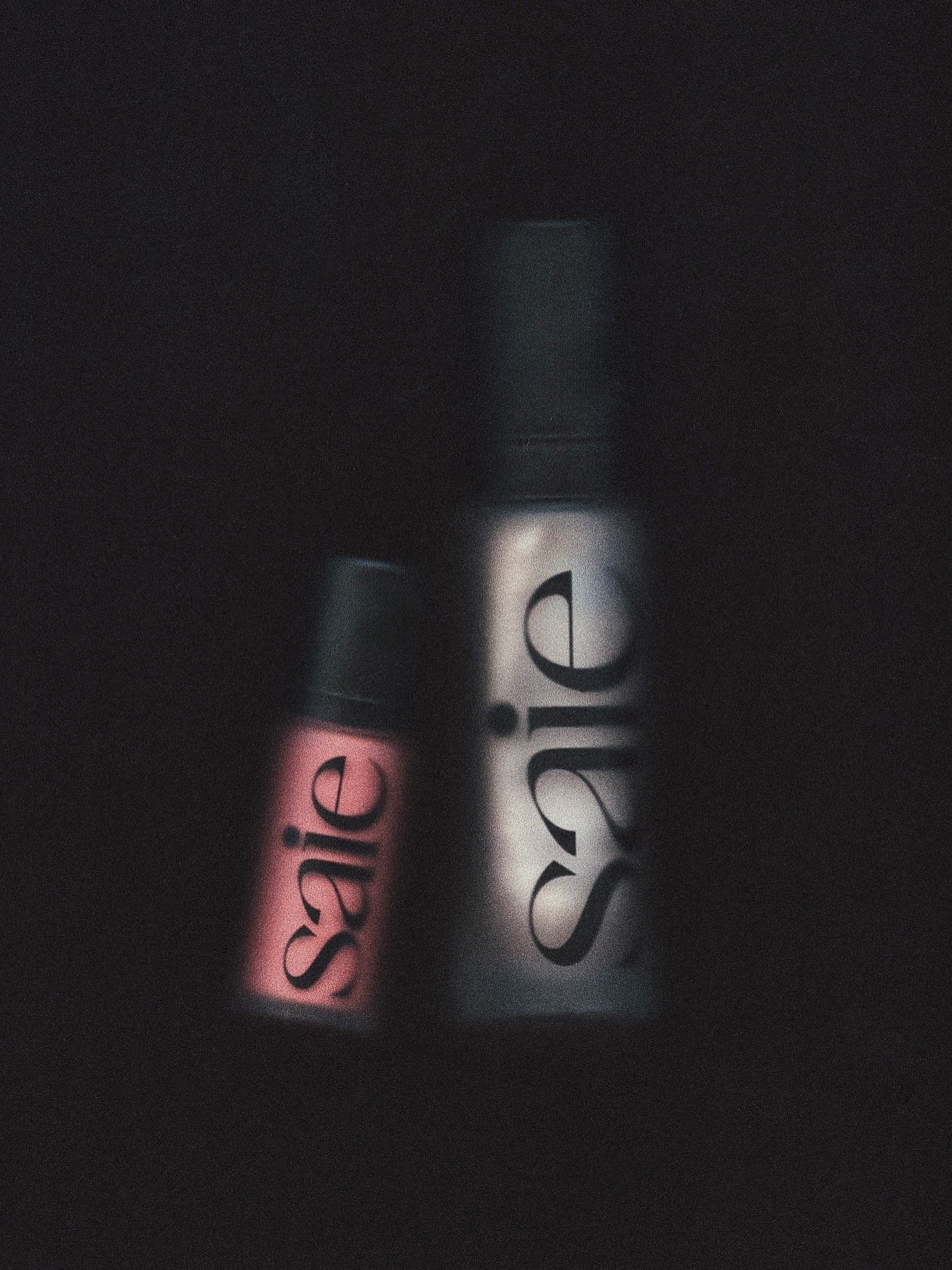 A close-up of two lip gloss tubes, one pink and one clear, with the brands' logos visible in a dimly lit setting.