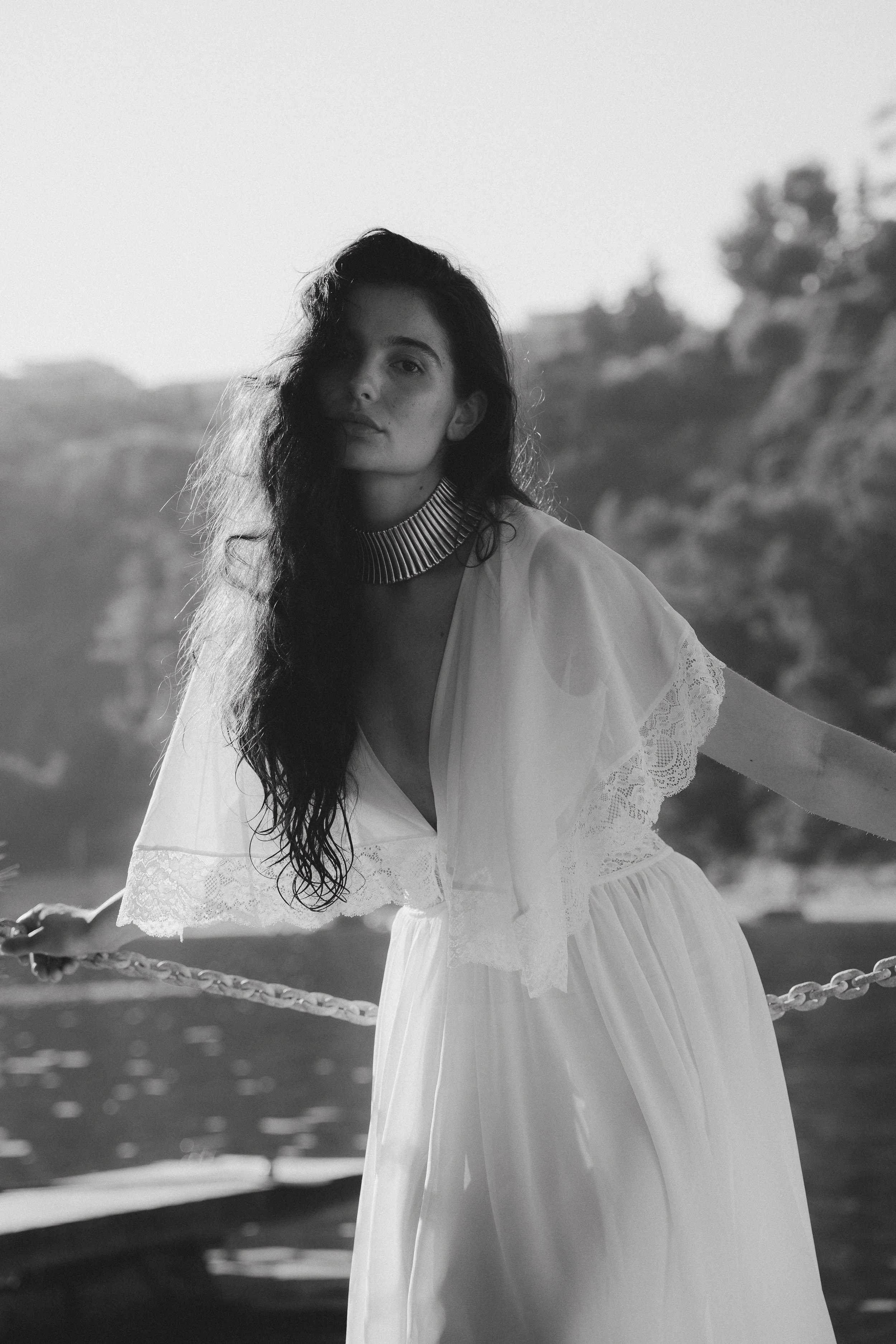 Black and white photo of a woman with long dark hair, wearing a white dress with lace details and a metallic choker, standing outdoors near water with blurred trees in the background.