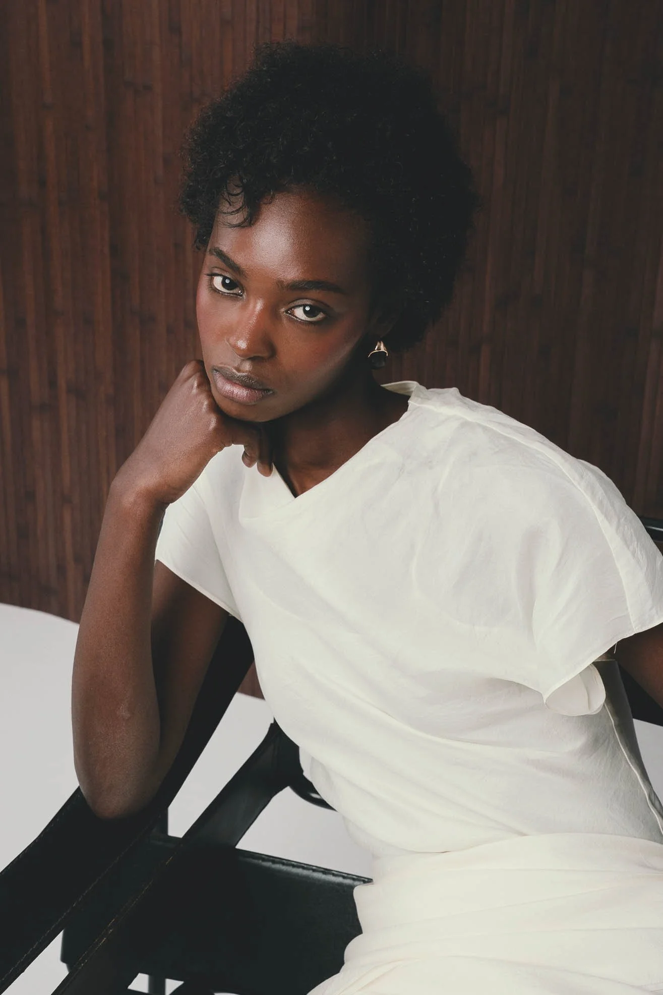 Portrait of a woman with dark skin with short curly hair, wearing a white top, seated on a black chair, resting her chin on her hand against a wooden background.