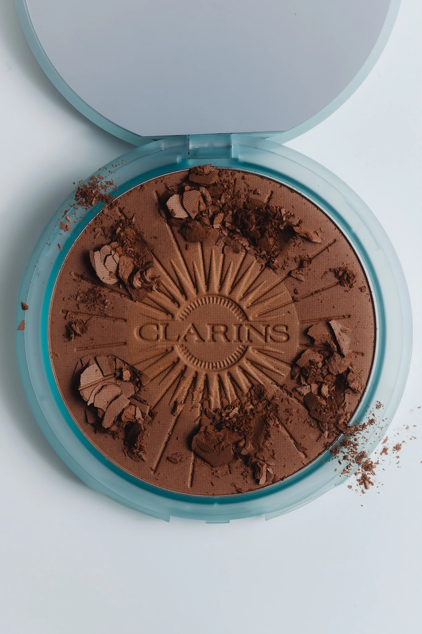 A round makeup compact opened to reveal pressed brown powder, with some broken pieces and crumbs on the surface and spreading out around it, with the brand name 'CLARINS' embossed in the center.