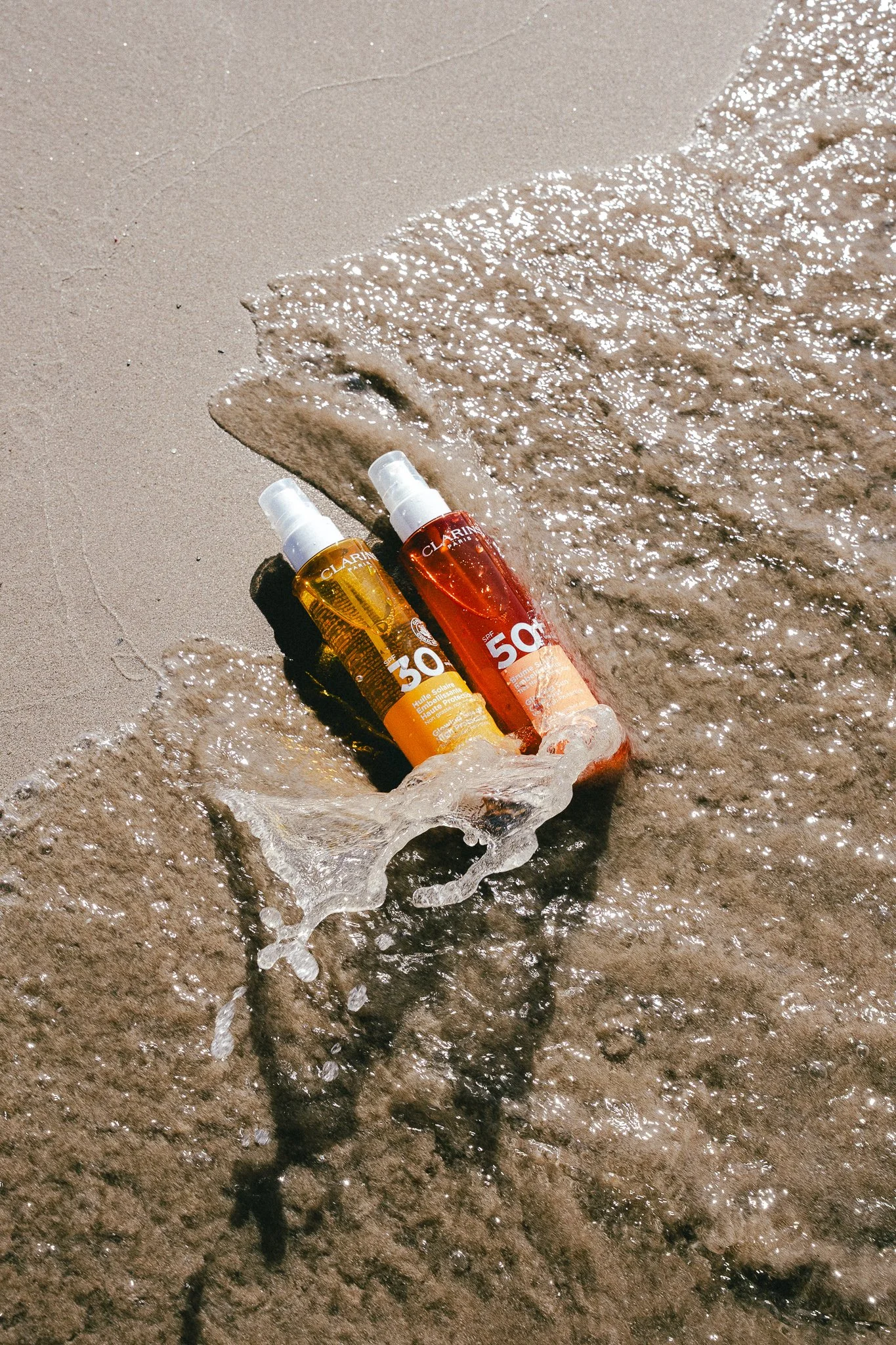 Two spray bottles of sunscreen, one with SPF 30 and yellow label and the other with SPF 50 and red label, lying in ice cube tray on sandy beach near the shoreline with gentle waves.