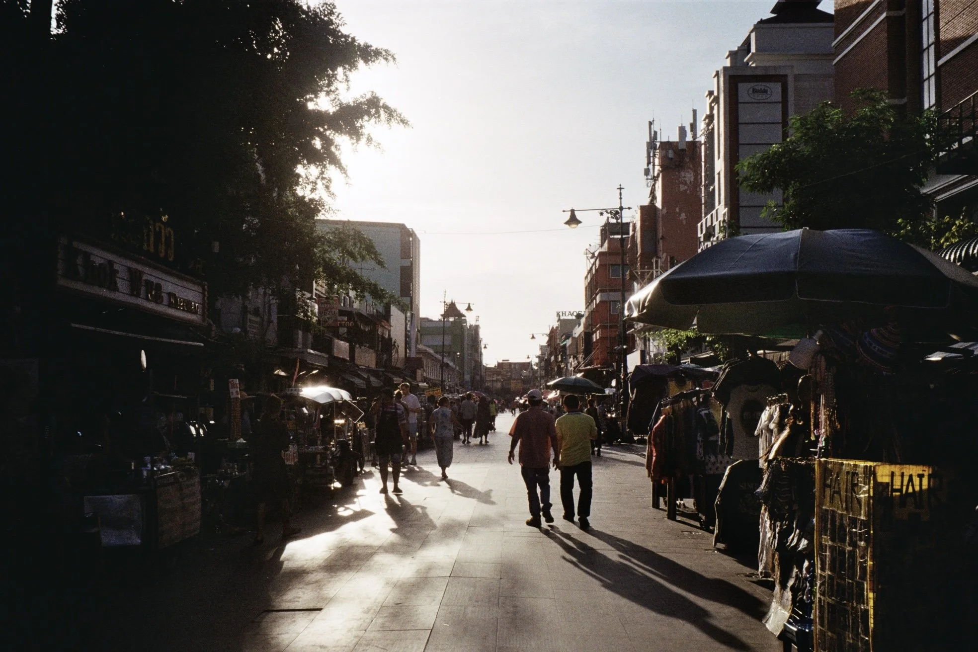 Crowded outdoor market street in an urban area during late afternoon or early evening with people walking, vendor stalls, umbrellas, and tall buildings in the background.