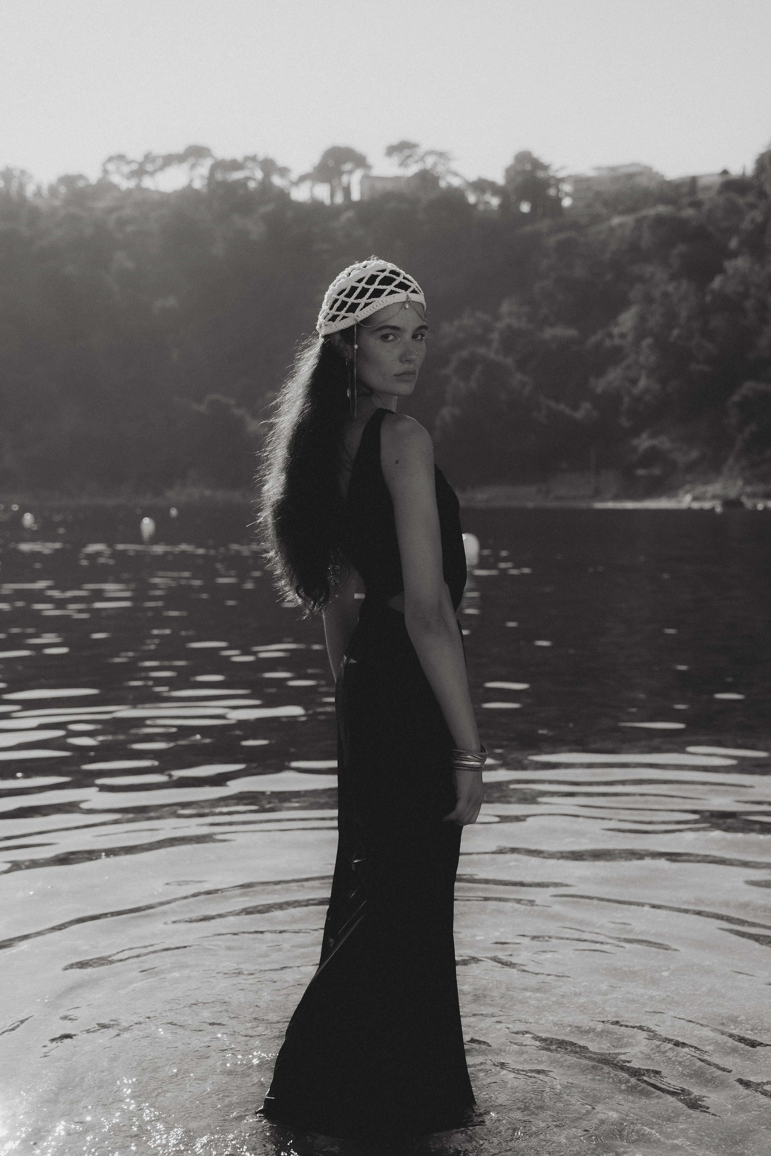 A woman with long hair and a decorative hat standing in water at the beach, with a hilly landscape and trees in the background. Black and white photo.