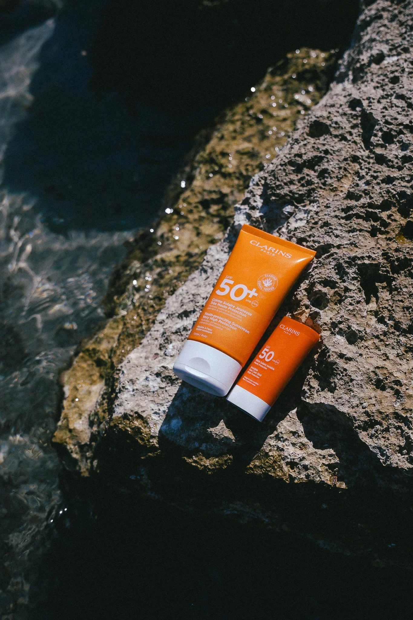 Two orange bottles of Clarins sunscreen with SPF 50+ resting on a rocky surface near water, sunlight reflecting off the wet rocks.