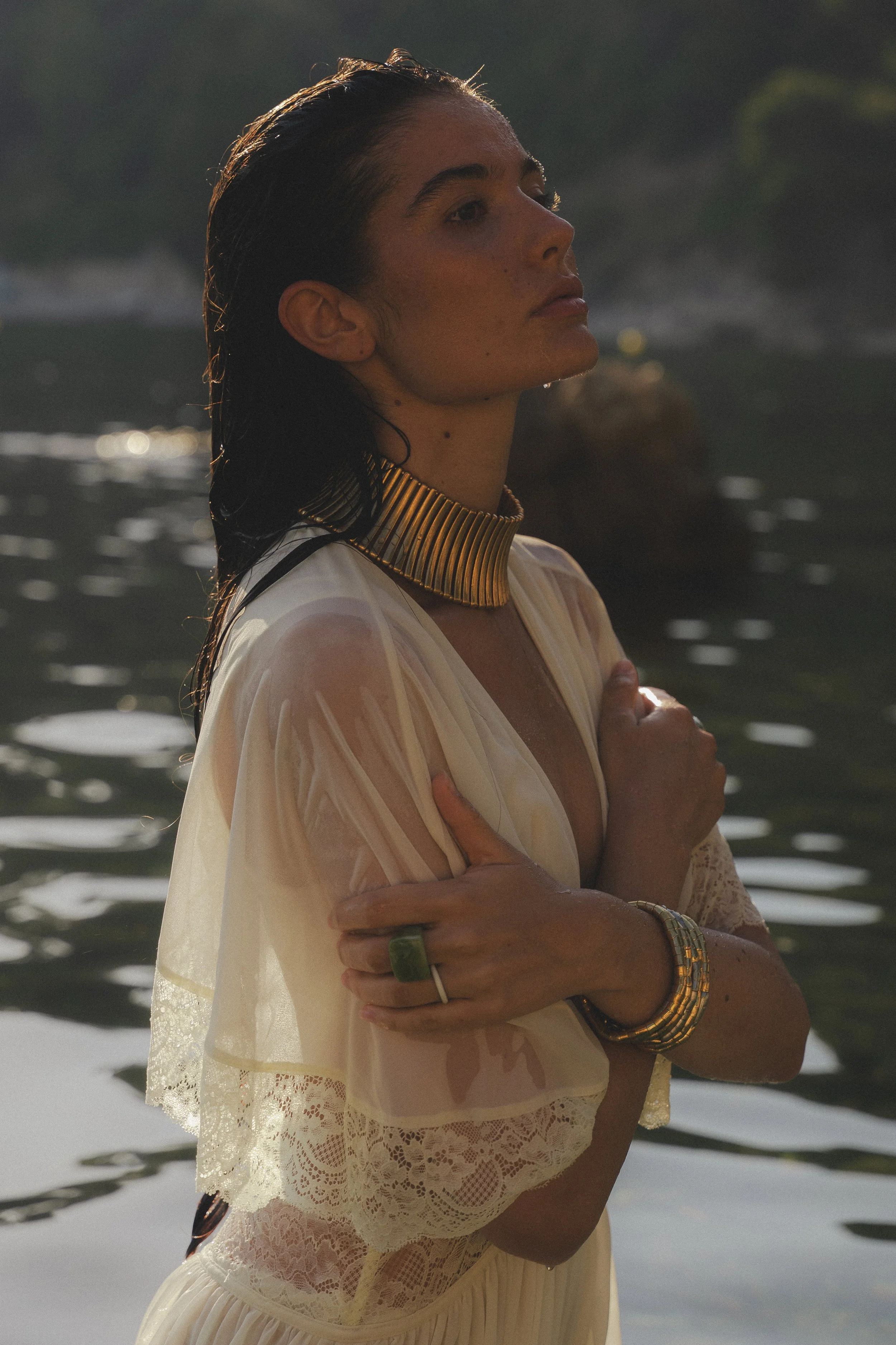 A woman with wet hair standing in water during sunset or sunrise, wearing a cream-colored dress with lace details, and gold jewelry including choker, bangles, and rings.