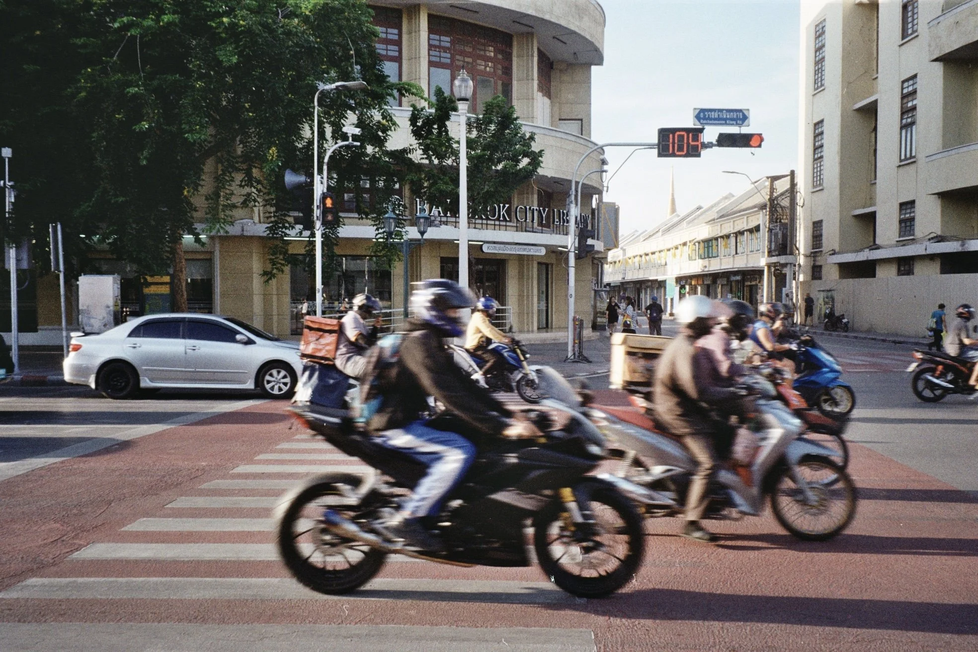 A busy city street with numerous motorcyclists riding through a crosswalk, cars, and pedestrians in the background, with a building sign reading 'Bangkok City Library' and traffic signals visible.