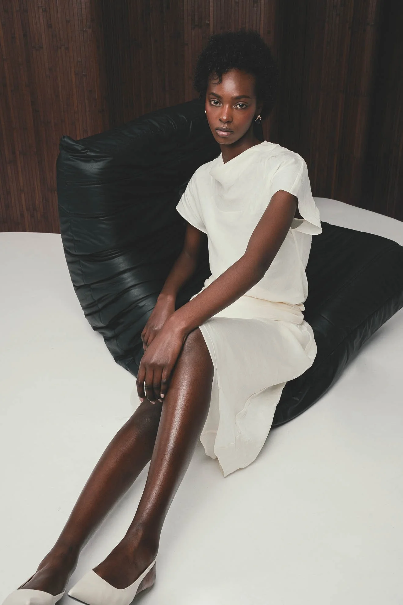 A woman with dark skin sitting on a large black bean bag chair with a wooden wall behind her. She is wearing a white dress and white high heels, with her legs crossed.