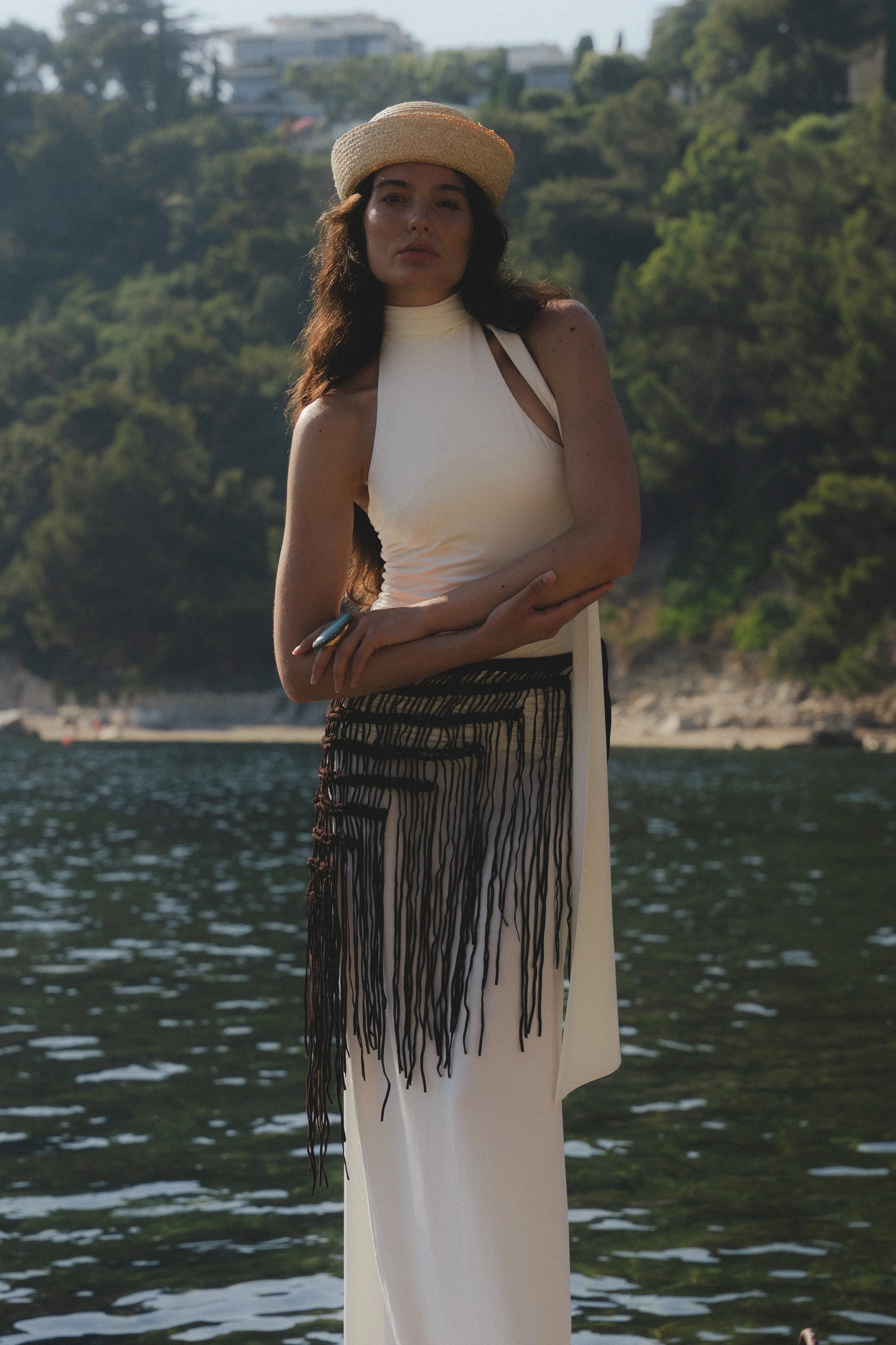 A woman in a white dress and a straw hat stands near a body of water with a forested hillside and buildings in the background.