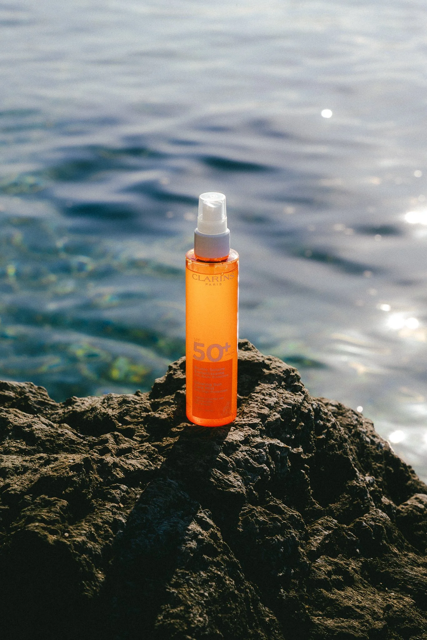 A bottle of sunscreen SPF 50+ is standing on a rugged rock near a body of water with sunlight reflecting off the surface.