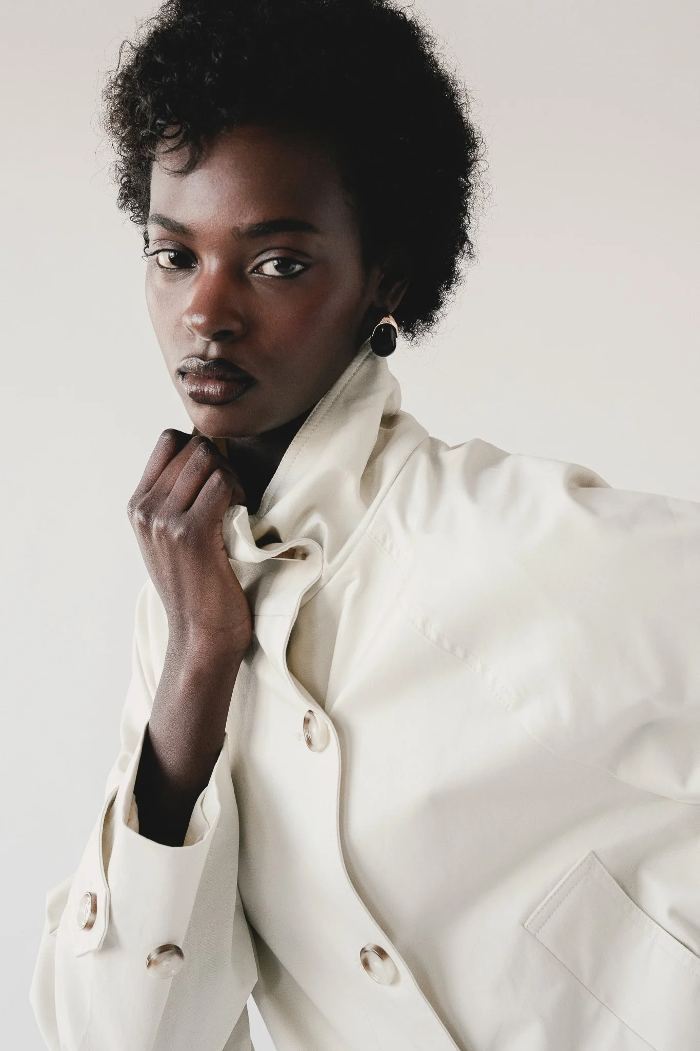 A portrait of a woman with dark skin and short, curly black hair. She is wearing a white trench coat with large buttons and black drop earrings. Her hand is gently resting on her collar, and she has a serious, confident expression against a plain whi