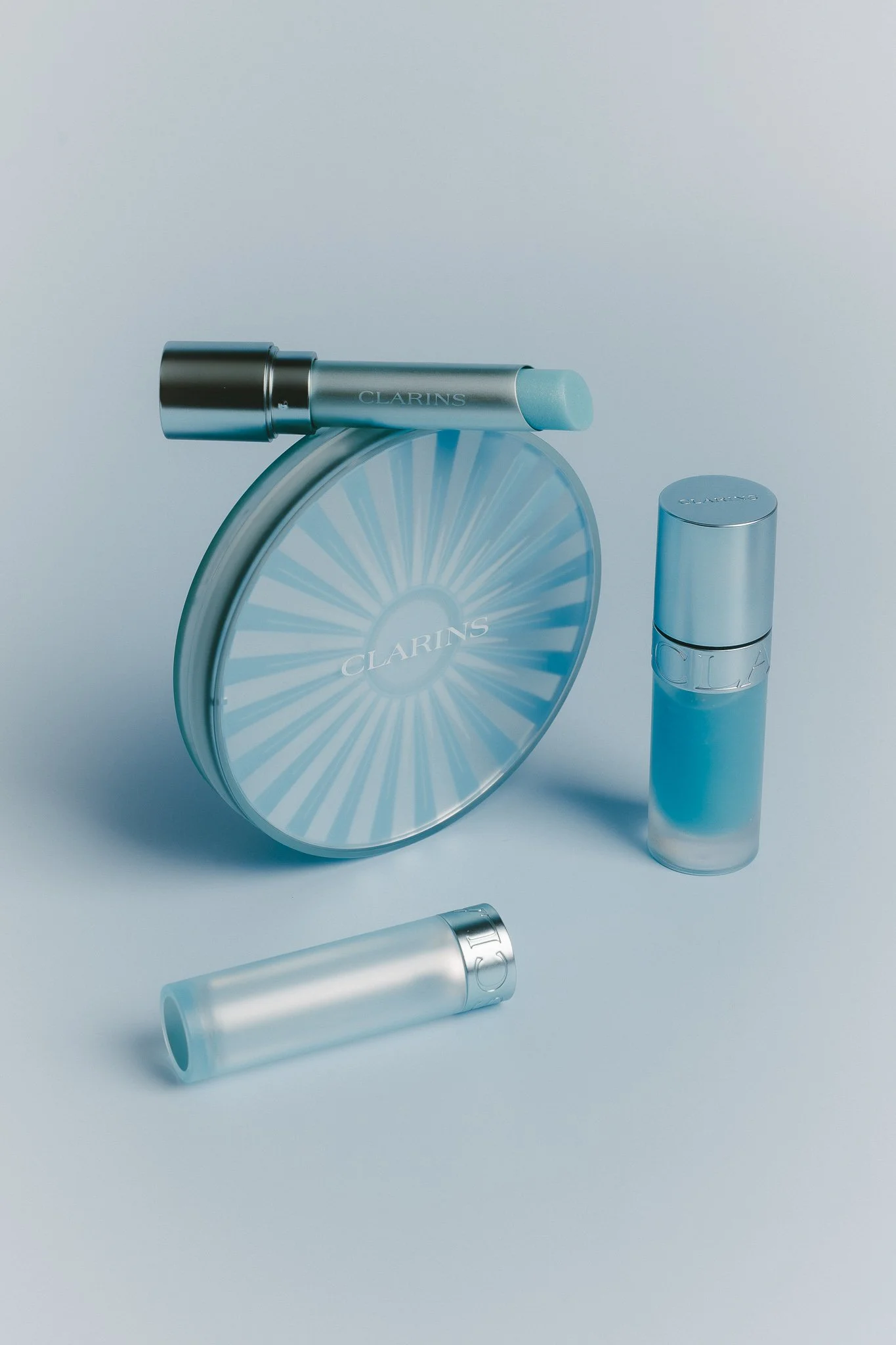 Various skincare products from Clarins, including cylindrical containers and a compact with a radial design, set against a light background.