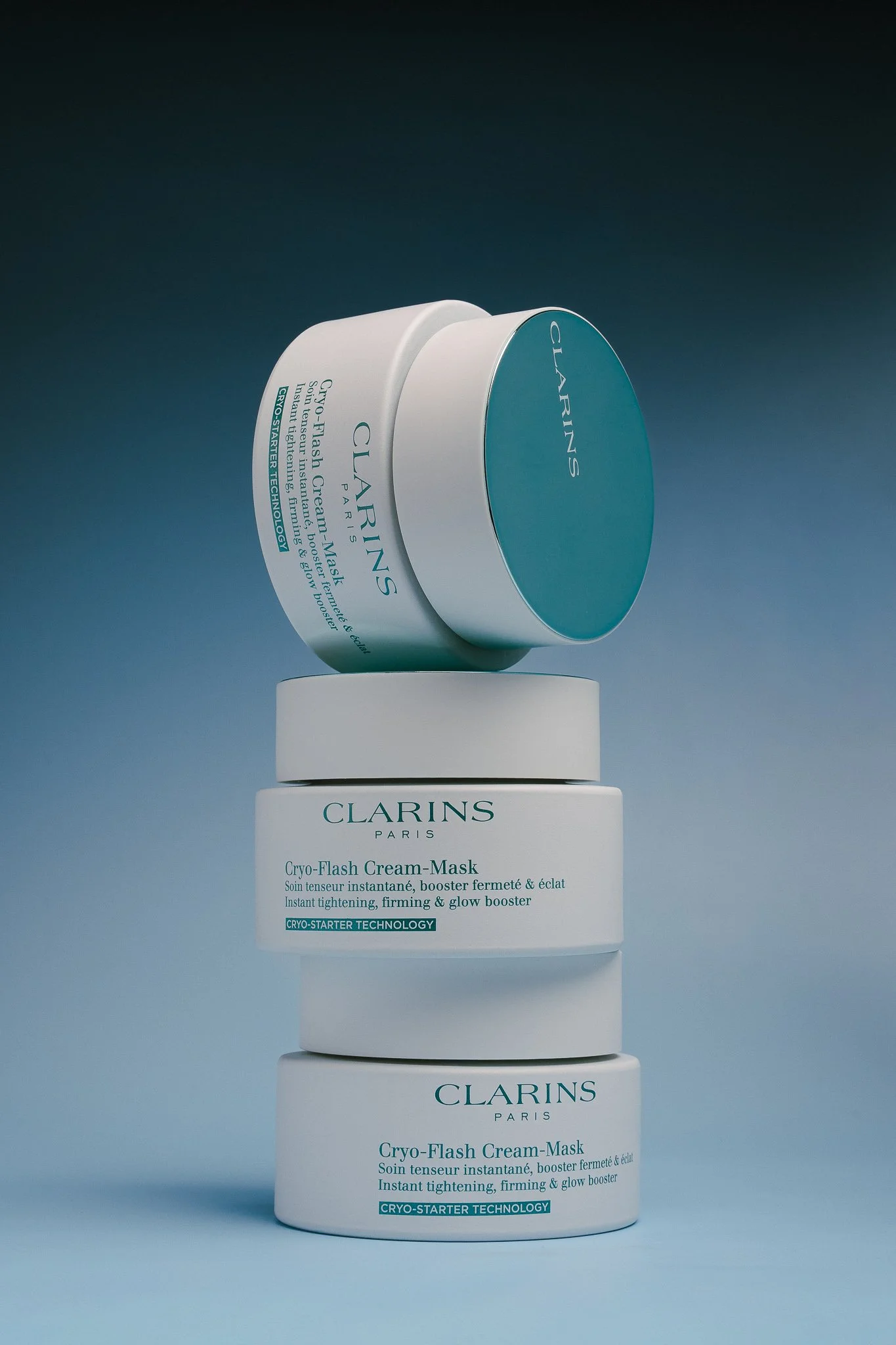 Stacked containers of Clarins Cryo-Flash Cream-Mask with blue lids, labeled as instant tightening, firming, and glow booster, against a gradient background.