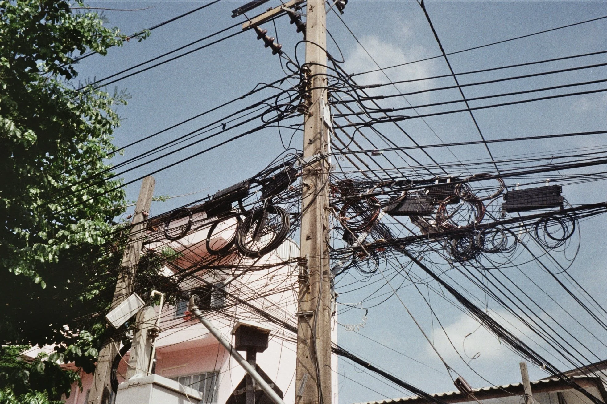 A utility pole with many tangled and disorganized electrical wires and components against a blue sky and some green leaves on the side.