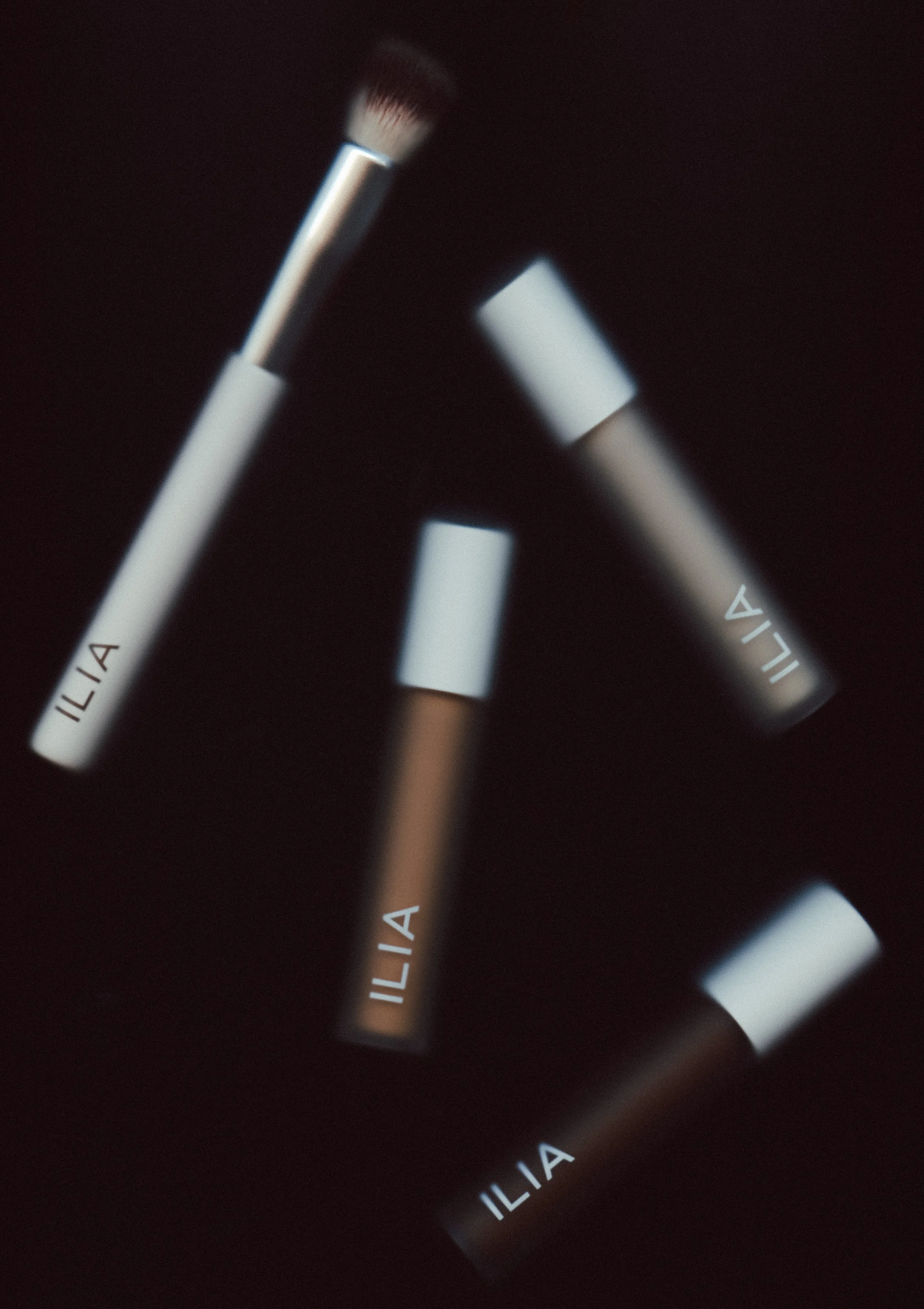 Close-up of illuminated perfume or cosmetic bottles with blurred white tops, with partially visible text "ILIA" on dark background.