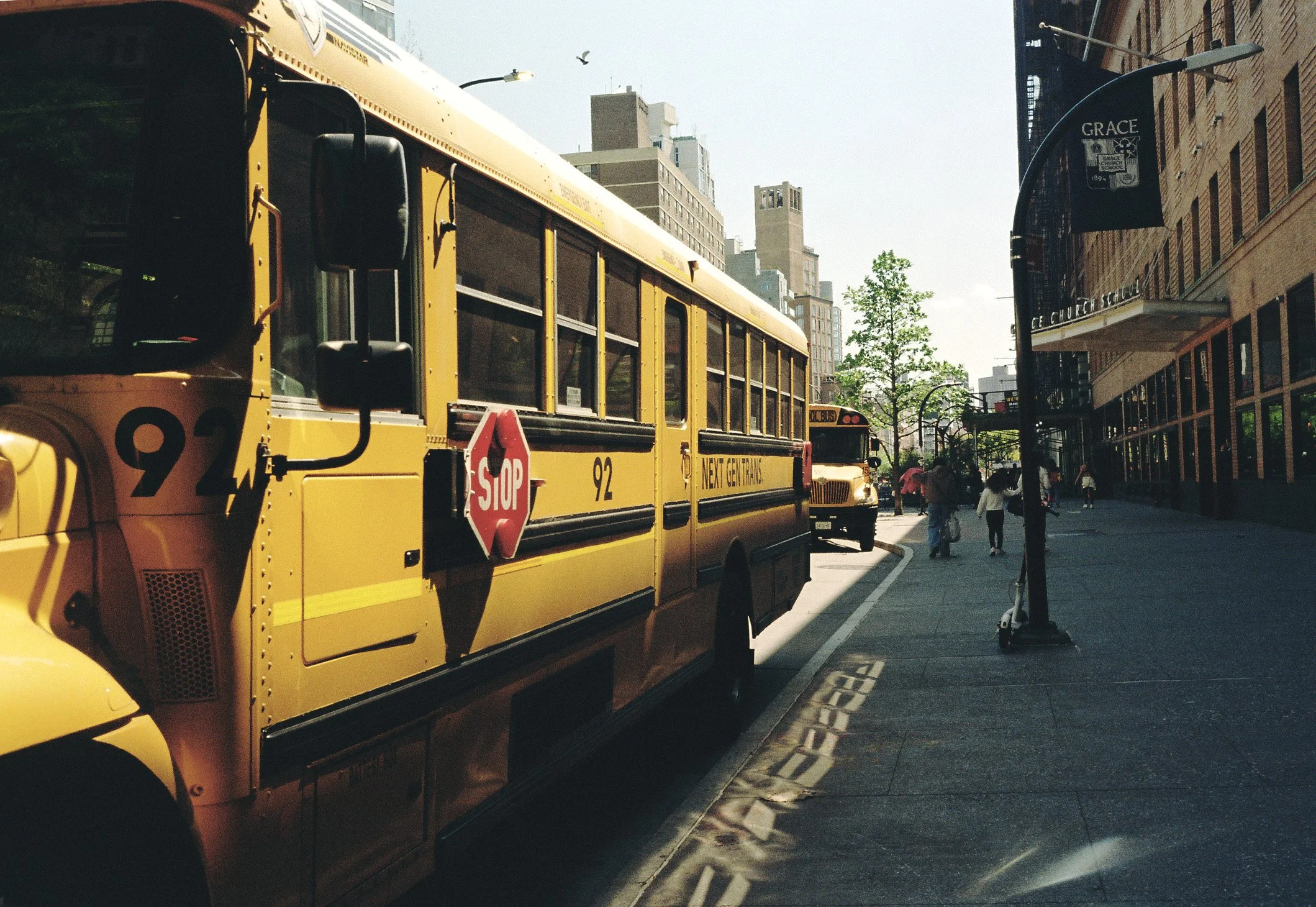 Yellow school bus parked on city street with pedestrians walking nearby, storefronts and buildings in the background, and a tree in the distance.
