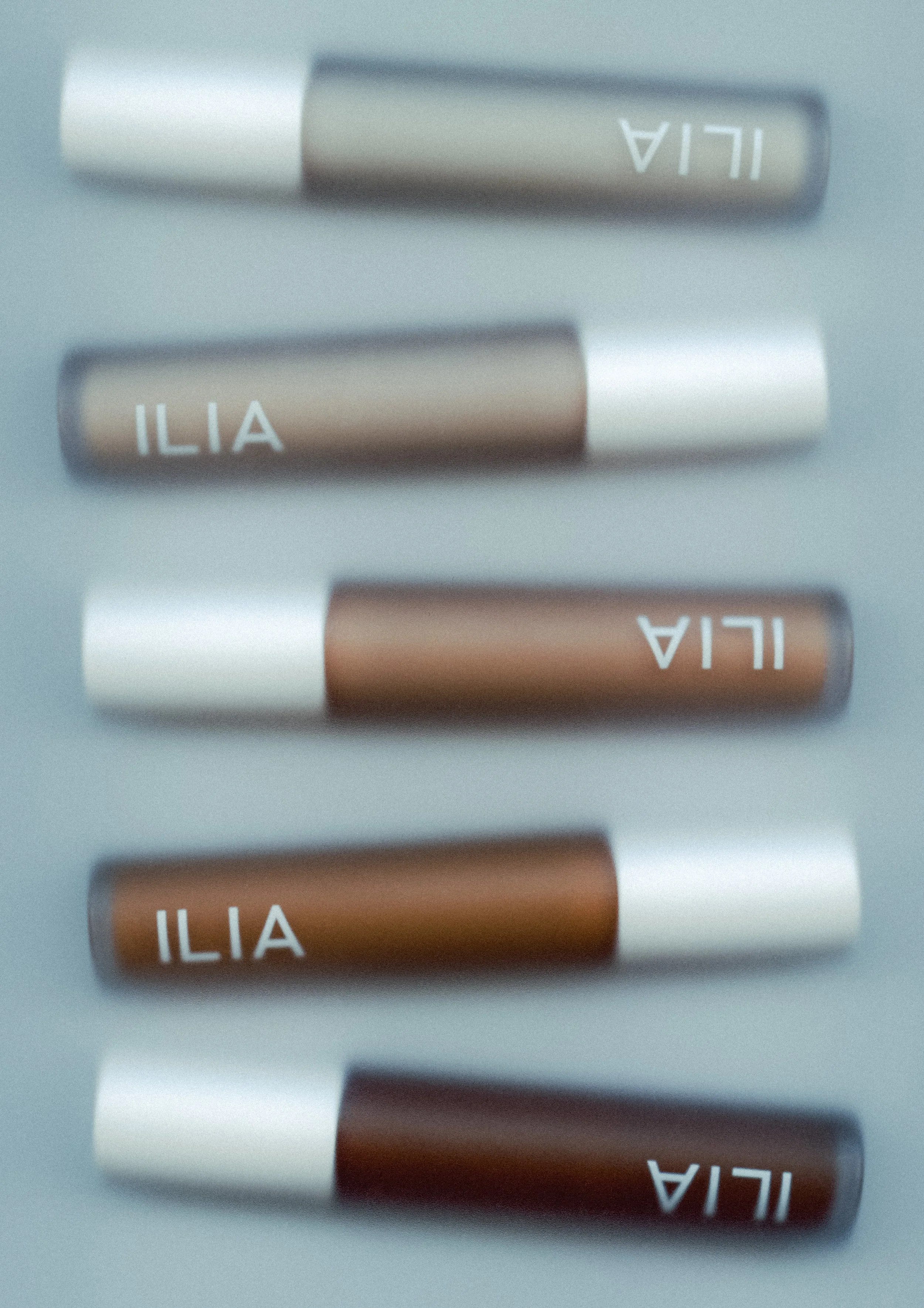 Close-up of three makeup lipstick tubes with metallic caps, labeled 'ILIA' on brown tubes and 'ILA' on gray tubes, all lying on a light-colored surface.