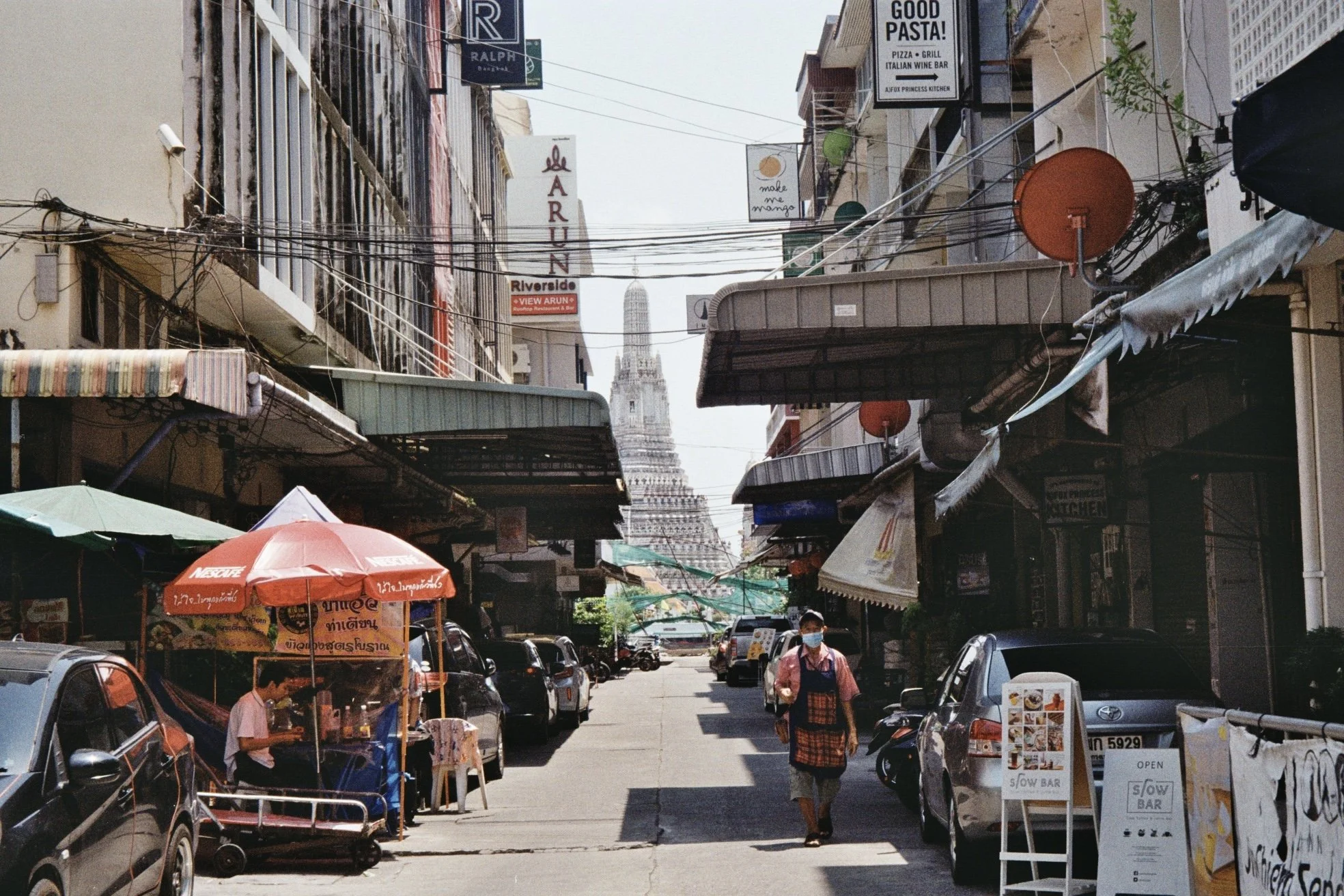 A busy street scene with parked cars, street vendors under umbrellas, and storefronts on both sides. The street is narrow, with signs in various languages, and a tall building or monument visible in the background.