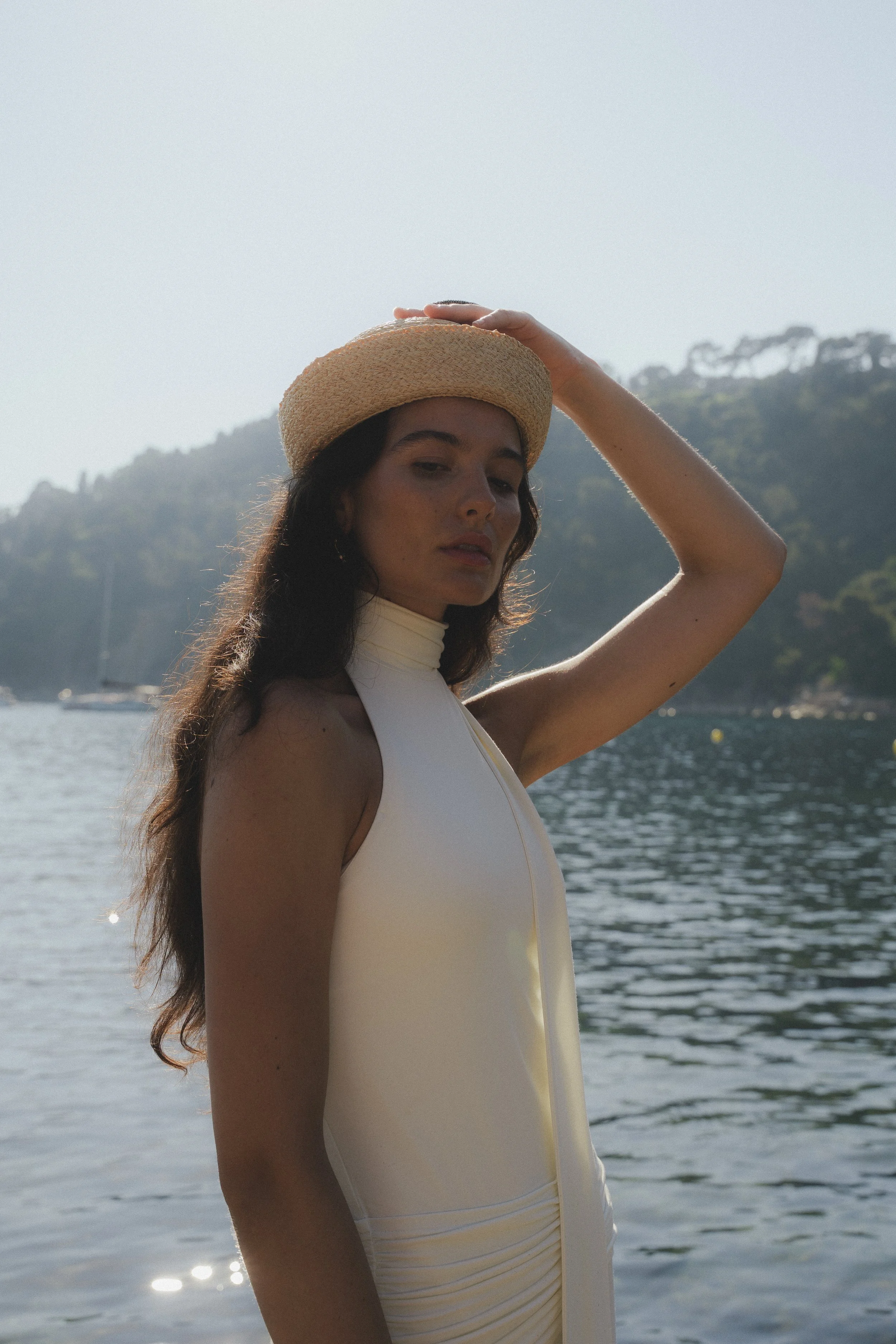 A woman wearing a beige sun hat and a sleeveless white dress standing by the water with hills and boats in the background.