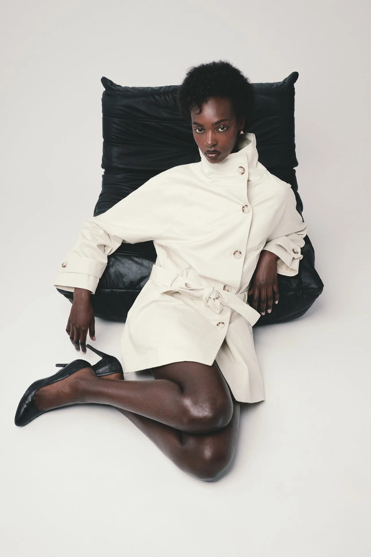 A woman with dark skin and curly hair is sitting on the floor, leaning against a large black cushion. She is wearing a cream-colored trench coat, black heels, and sheer black tights, with one leg stretched out and the other bent. Her expression is se