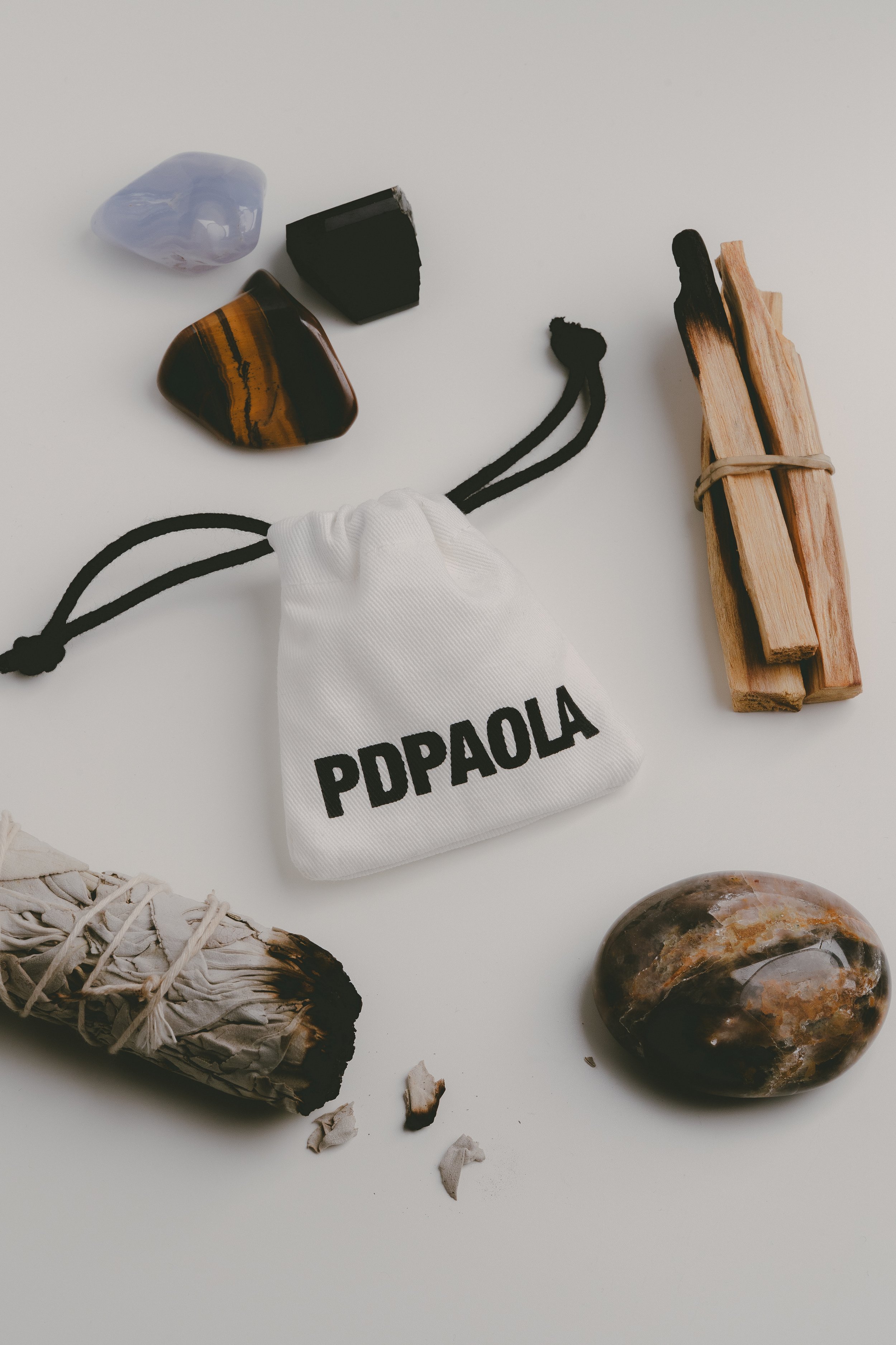A white cloth bag with black string pockets labeled 'PDPAOLA' containing a black stone, a purple stone, and a tiger's eye stone, on a white surface. Nearby are a bundle of sage, a polished stone, and a bundle of wooden sticks bound with rubber band.