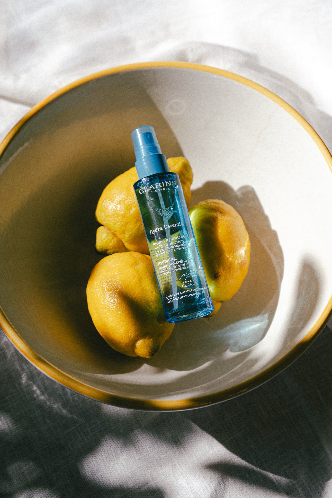 A bowl containing four yellow lemons with a blue spray bottle of Clarins Hydra-Essentiel in the center.