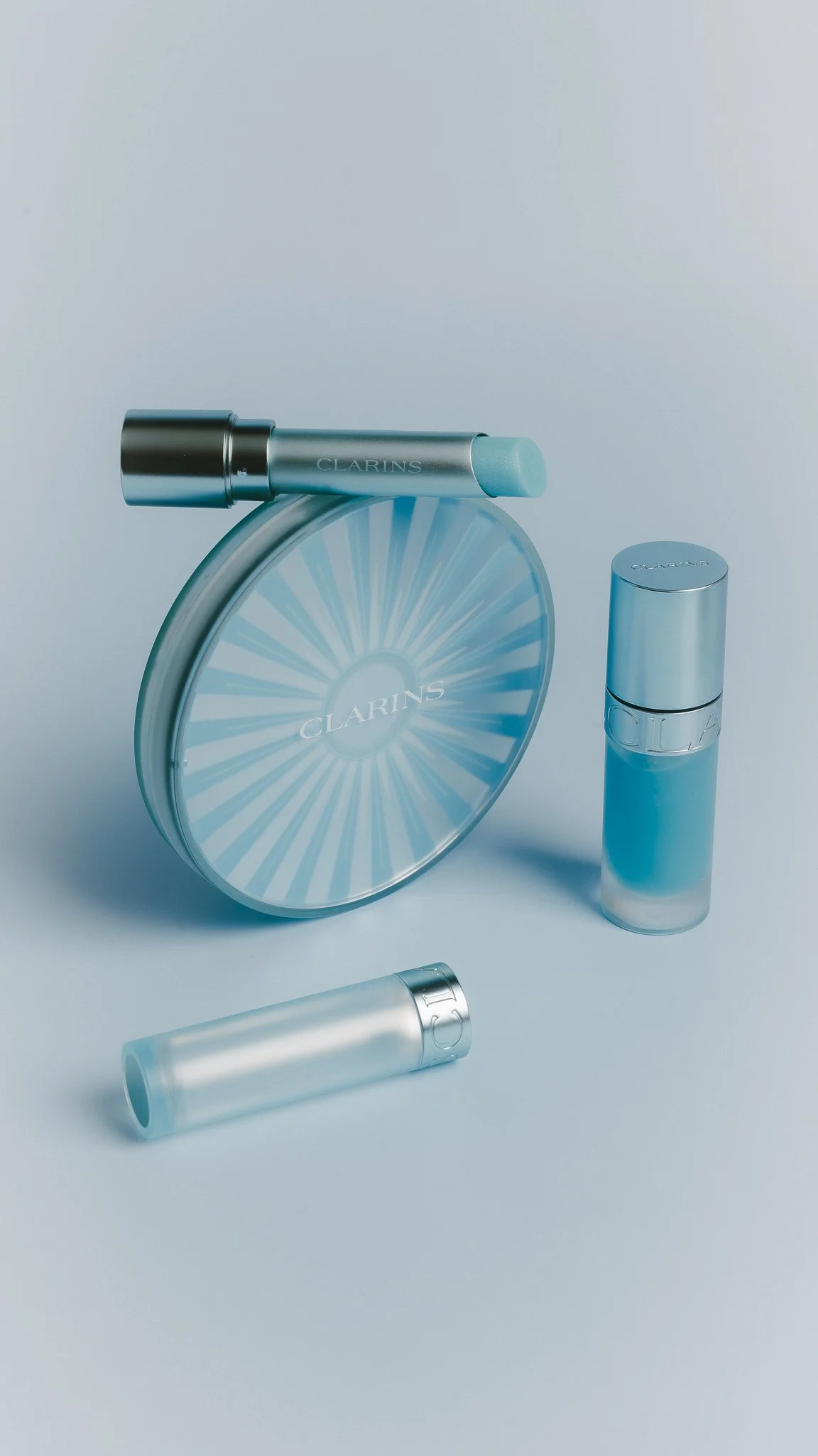 Collection of Clarins skincare products including a compact case, a stick, and several cylindrical bottles, all in a light blue and silver color scheme on a white background.