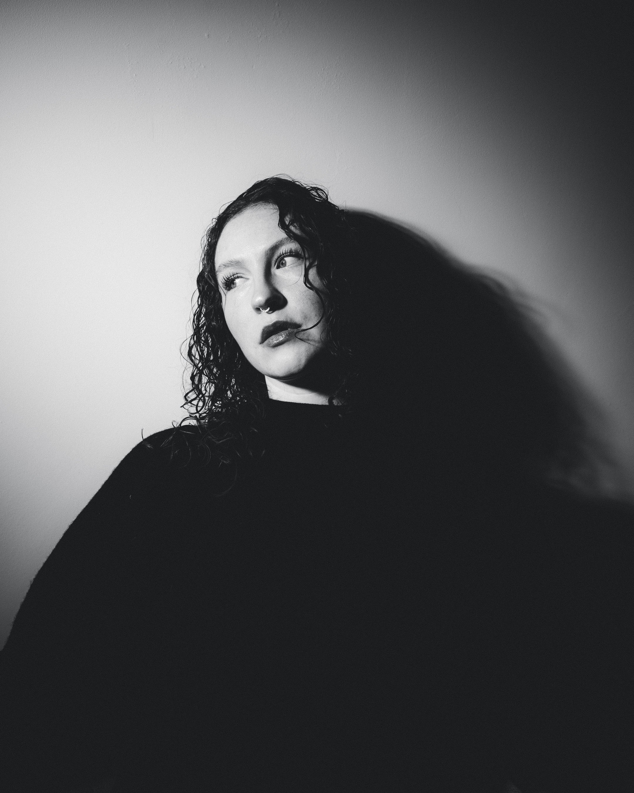 A black and white portrait of a woman with curly hair, looking to her right, with a septum piercing, dark lipstick, and wearing a dark top.