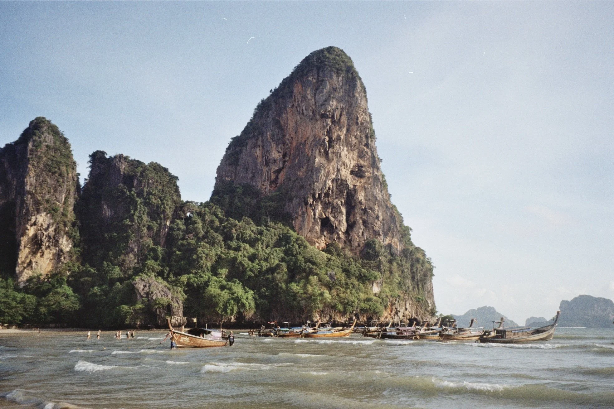A scenic coastal landscape featuring tall limestone cliffs covered in greenery, with boats docked by the shore and calm waters with gentle waves.