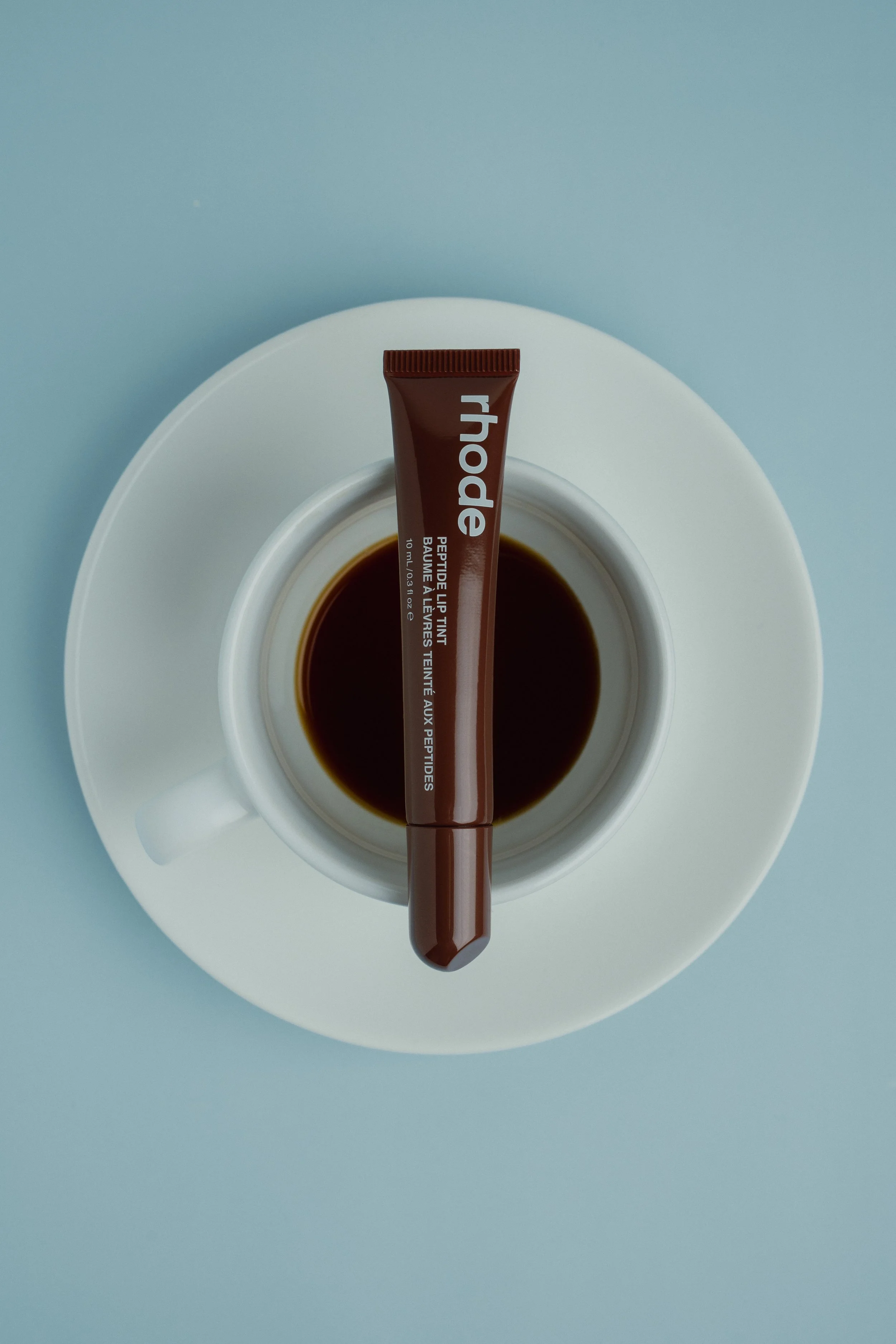 A brown tube of lipstick resting on the rim of a white coffee cup filled with black coffee. The coffee cup is placed on a white saucer, and the background is a light blue surface.