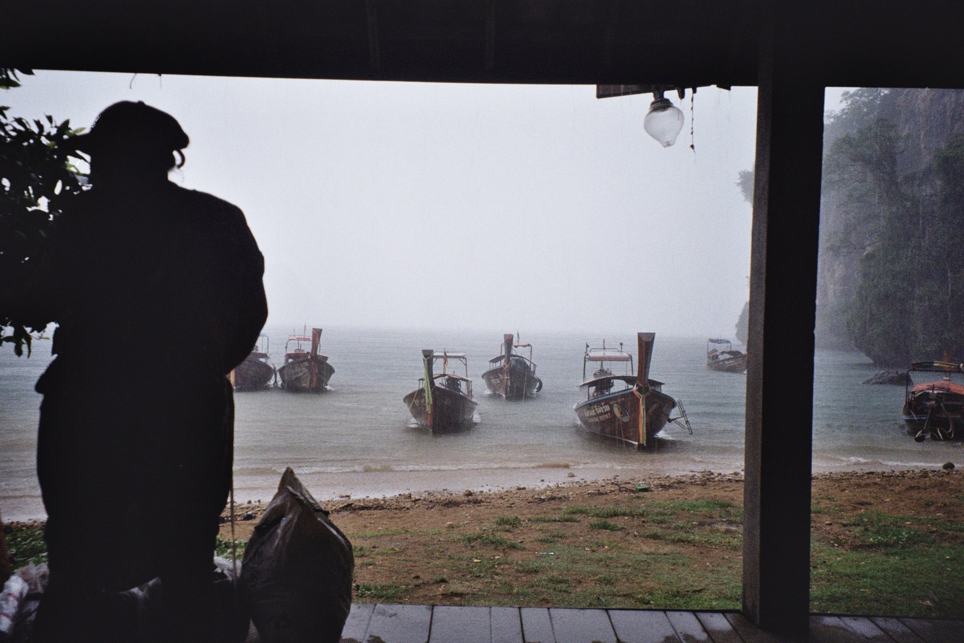 A person in silhouette standing under a gazebo, looking out at boats docked along the shore on a foggy day.