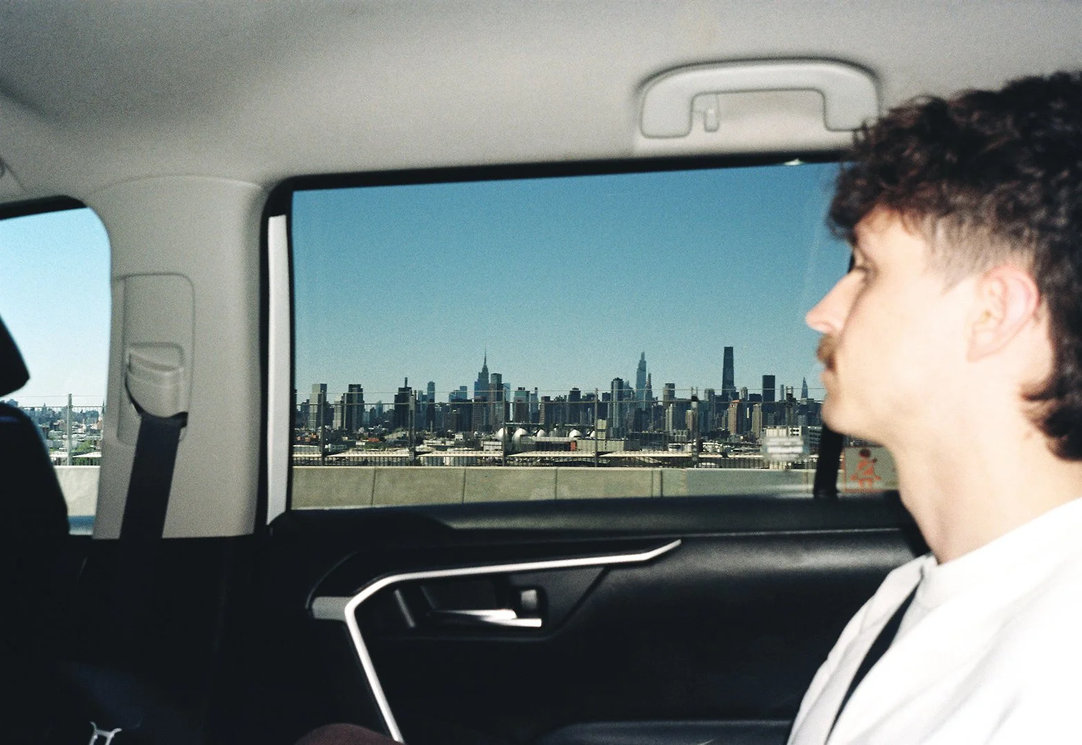 A man with brown hair and a mustache looking out the window of a vehicle at the New York City skyline in the distance.