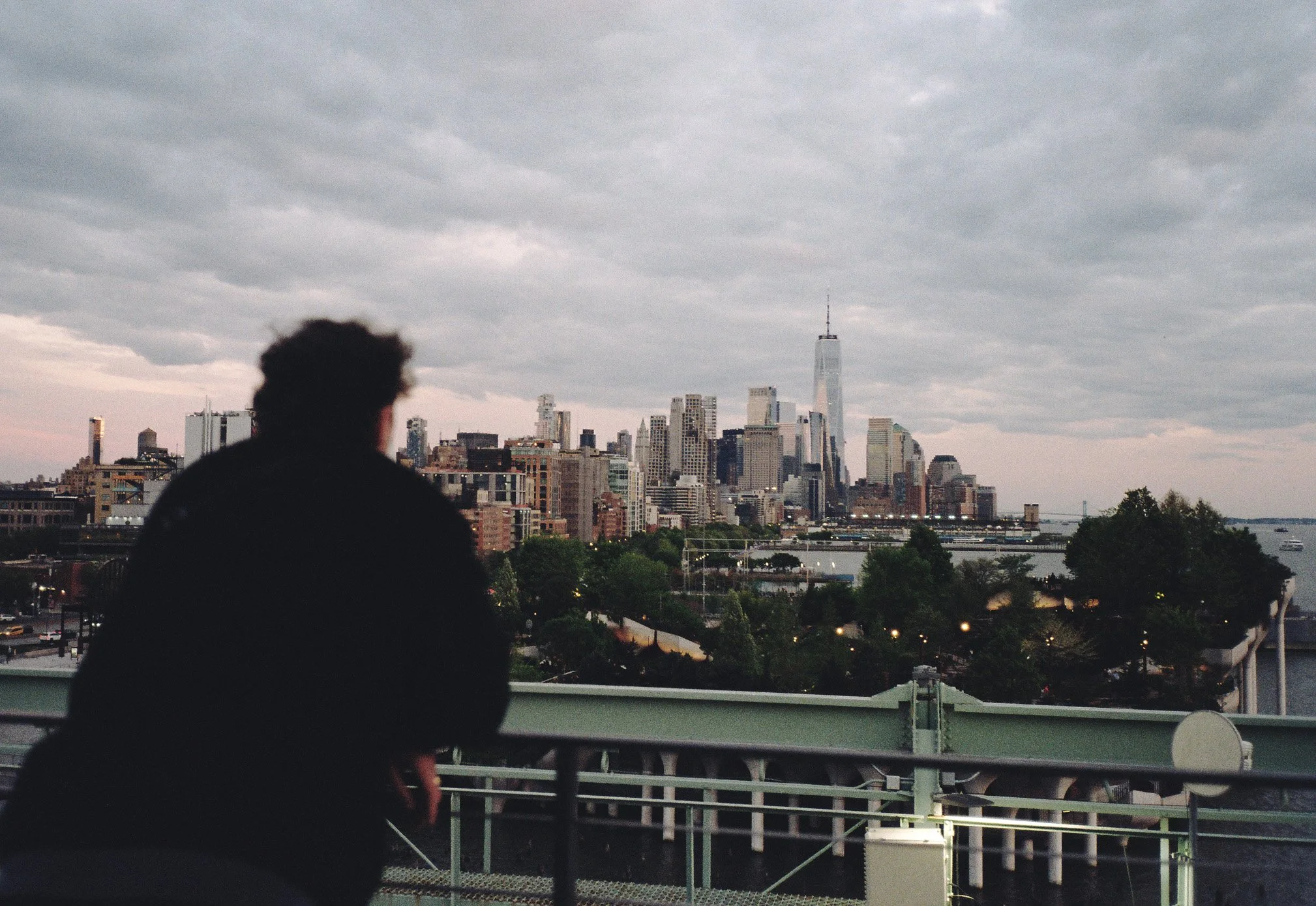 A person with short curly hair looking at the city skyline of New York City on a cloudy day from a bridge or balcony.