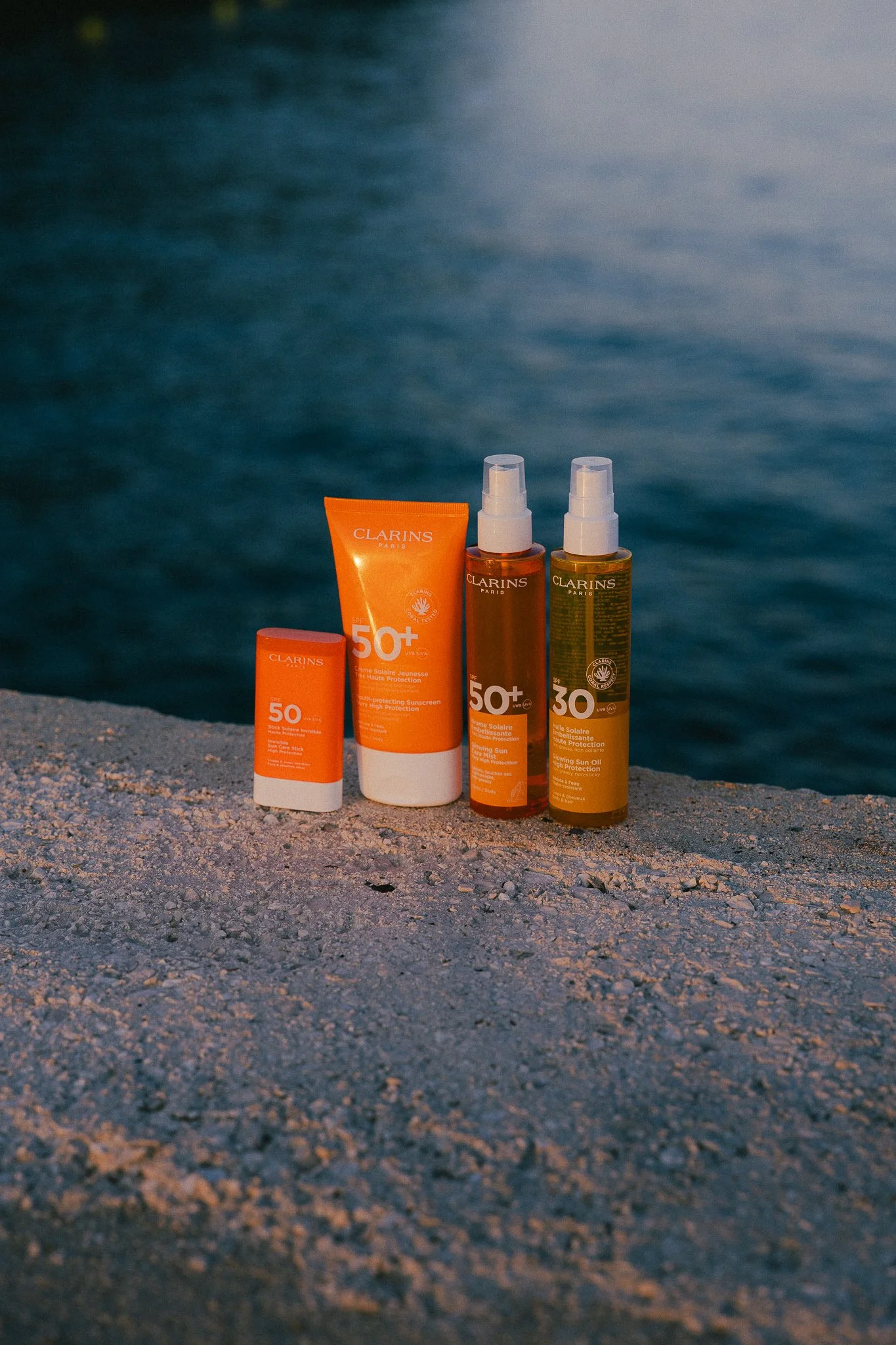 Four bottles of Clarins sunscreen products on a gravel surface near water. Two bottles have SPF 50+ and two have SPF 30, with the bottles featuring orange and green labels.