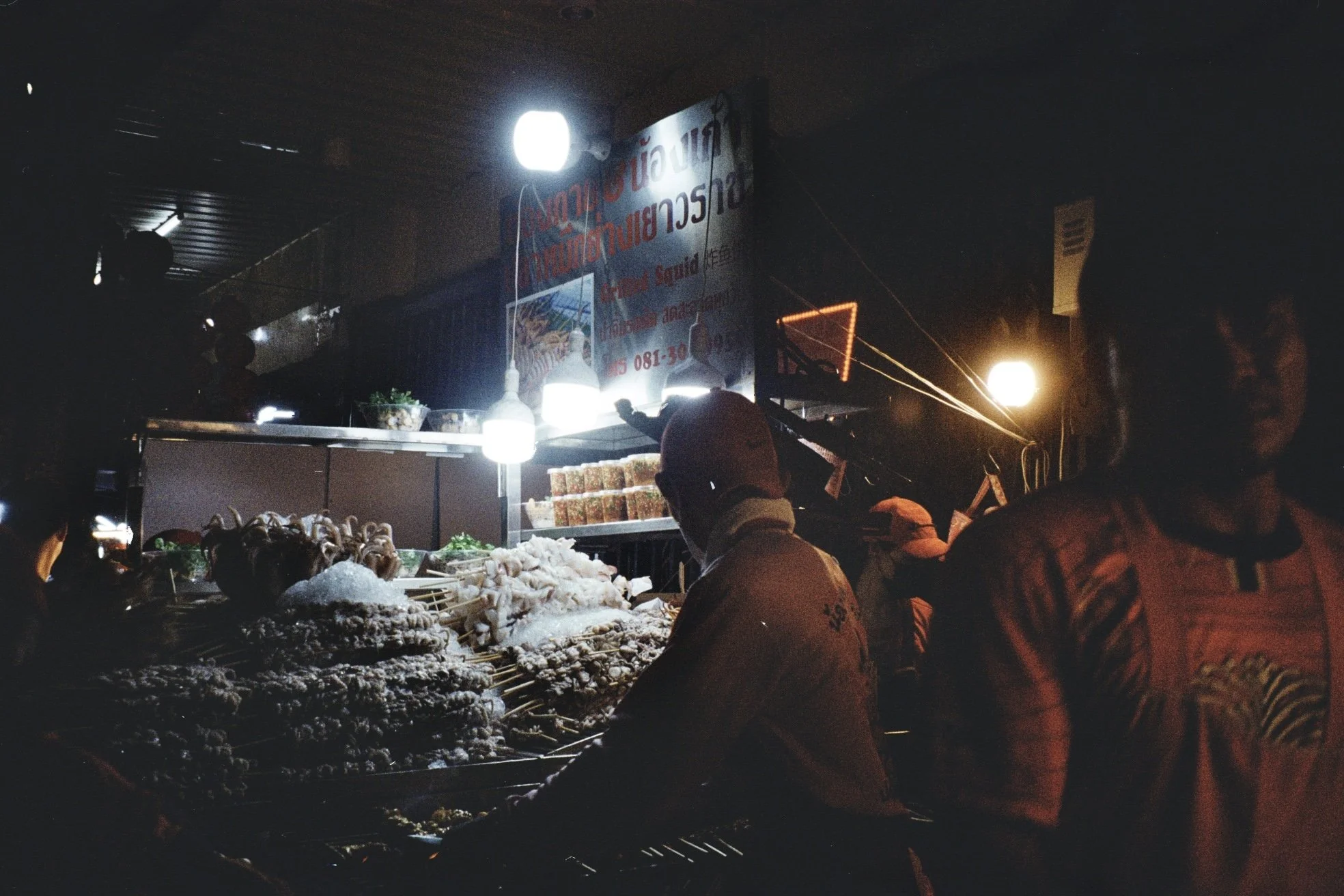 Night scene at a street food stall with bright lights, showing food items on display, including skewers and vegetables, with people working behind the counter and some customers in front, illuminated by overhead lights.