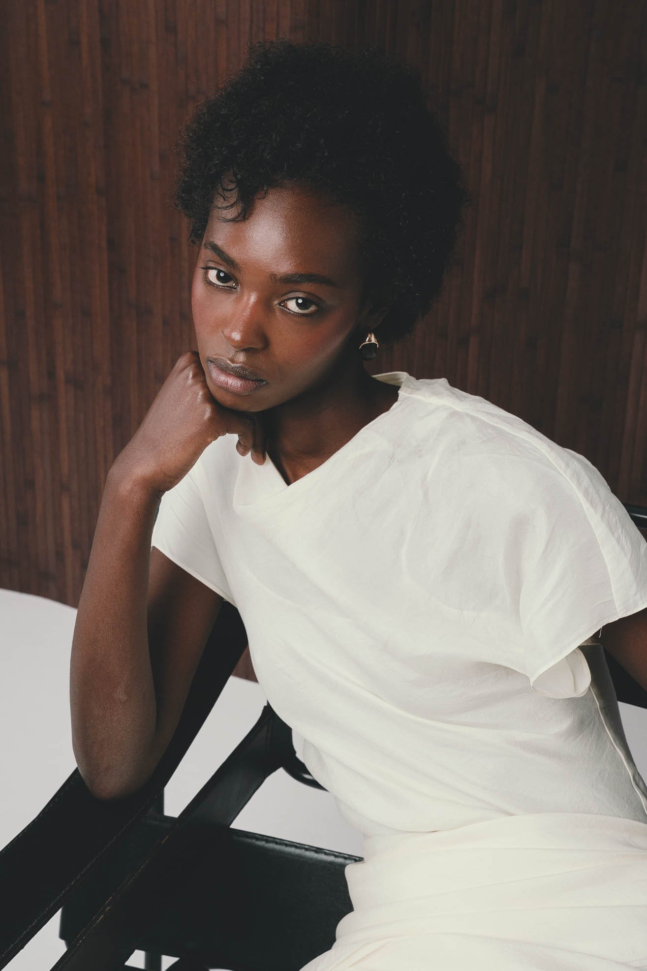 Portrait of a young woman with dark skin, short curly hair, wearing a white top, sitting on a black chair, with a wooden background.