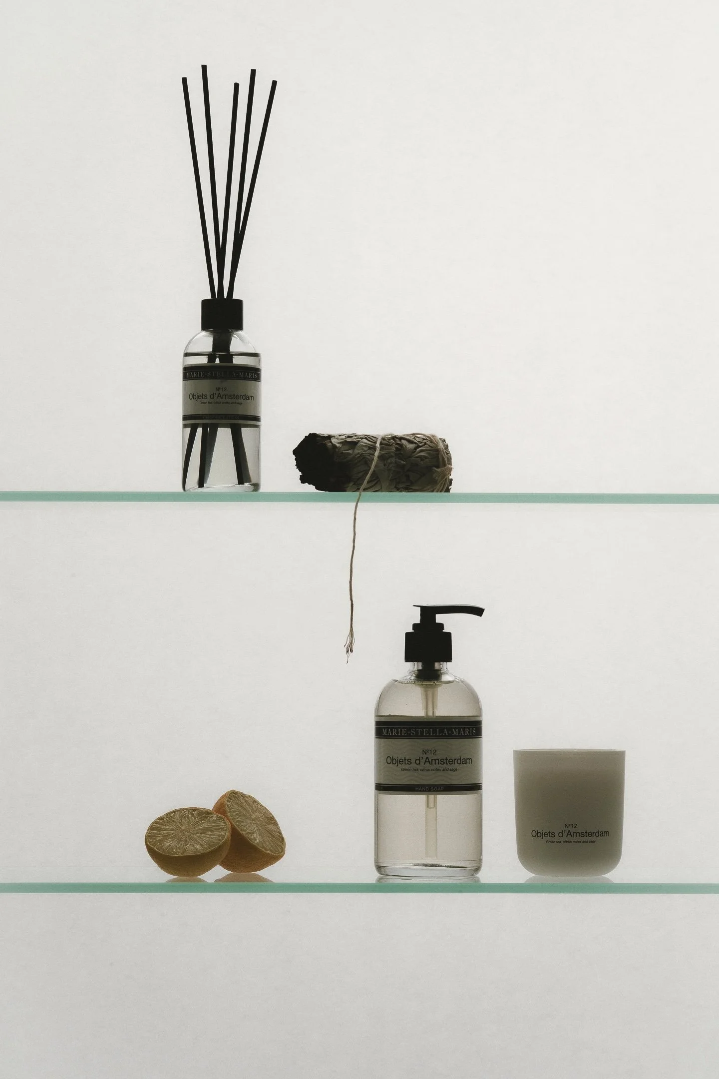 A minimalist display of personal care items and objects on a white background, including black bamboo diffuser, dried sage bundle, a bottle of hand soap, a white cup, and halved lemon slices.