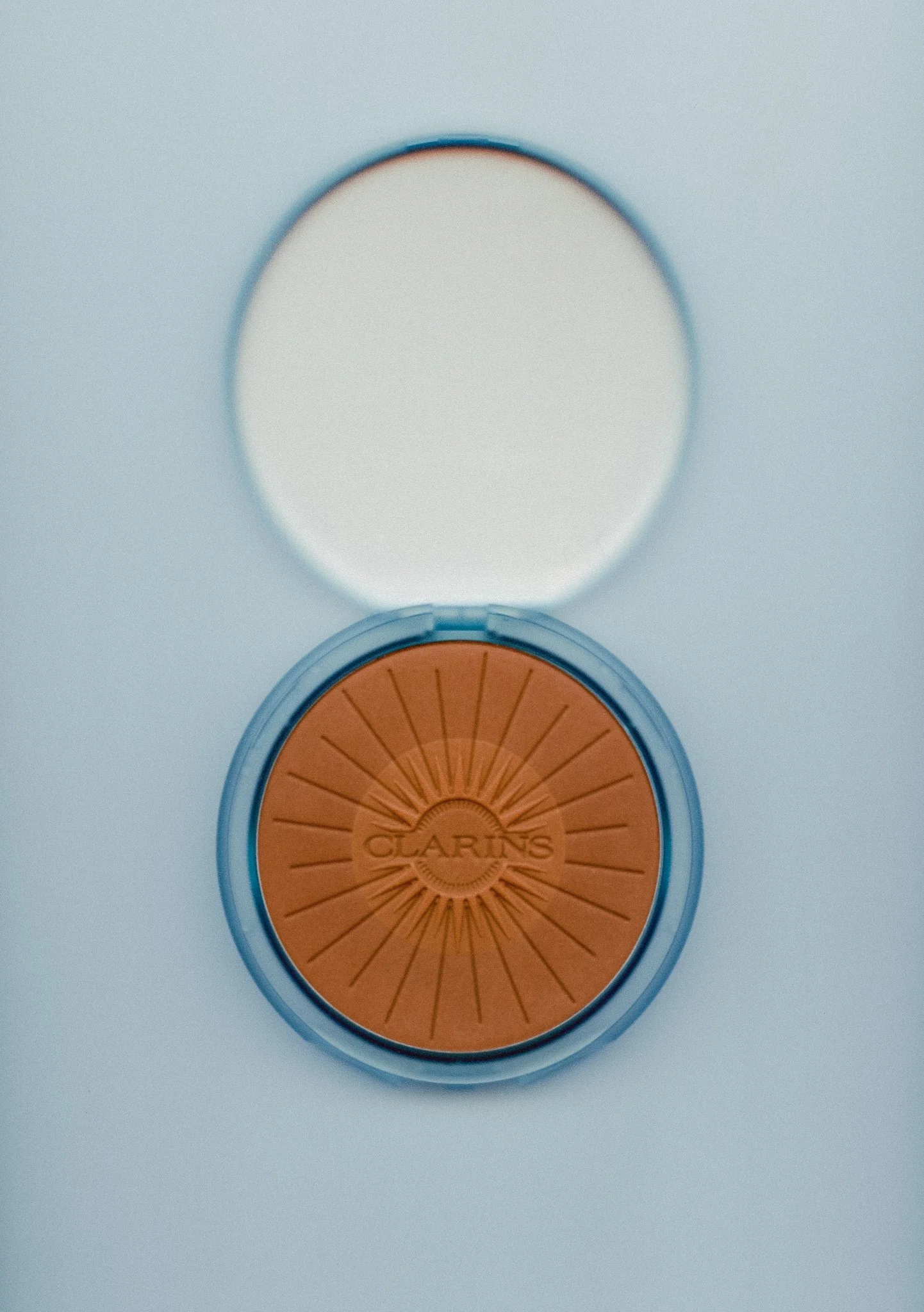 round orange makeup palette with a sunburst design and the word 'CLARINS' in the center, open with mirror reflecting the palette