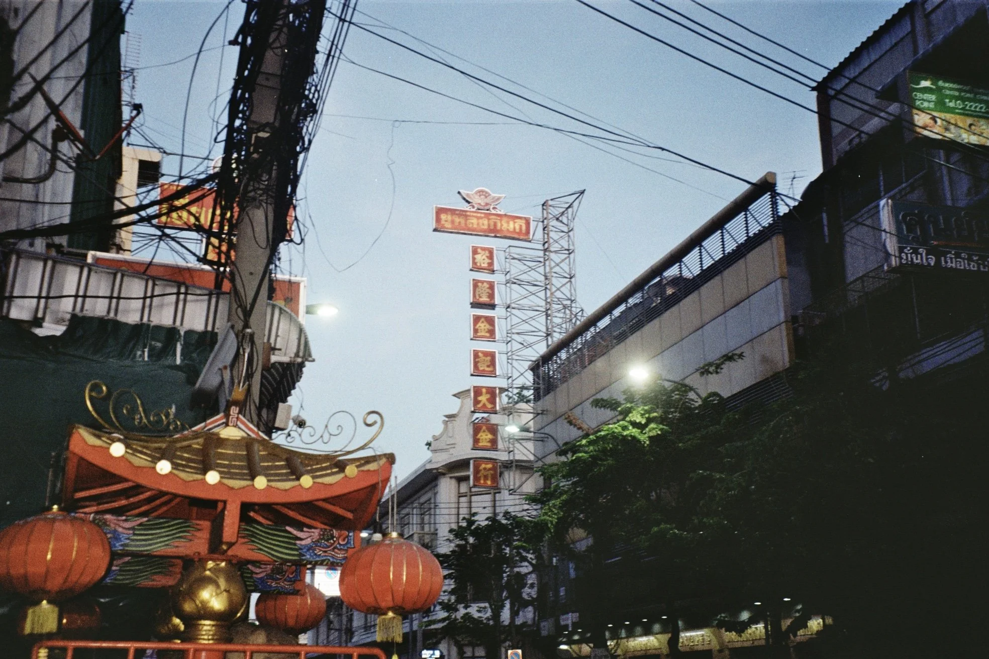 Street scene at dusk with signage in Thai, red lanterns, and overhead electrical wires.