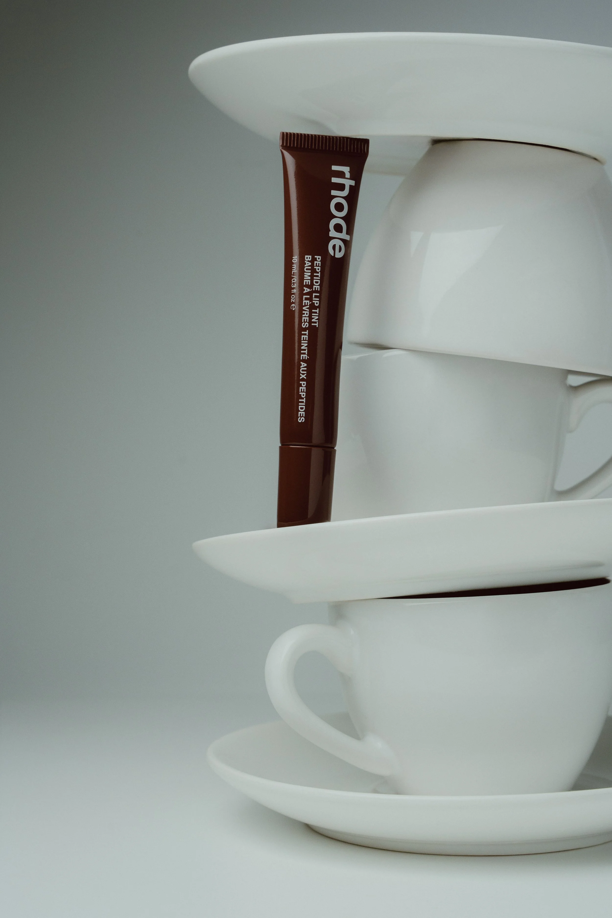 A white teapot with a white cup inside it, and a tube of lip tint lipstick hanging on the teapot's handle, on a white surface with a gray background.