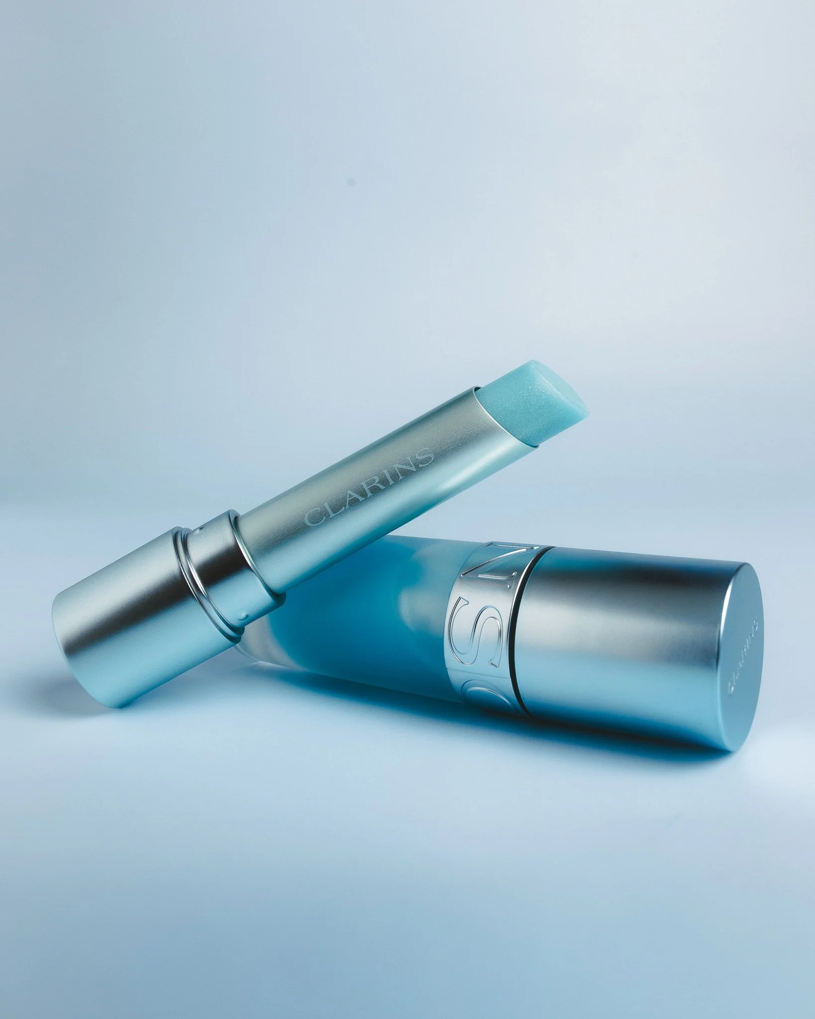 Close-up of three Clarin's lip balms in pastel shades of blue, silver, and gray, lying on a light blue background.