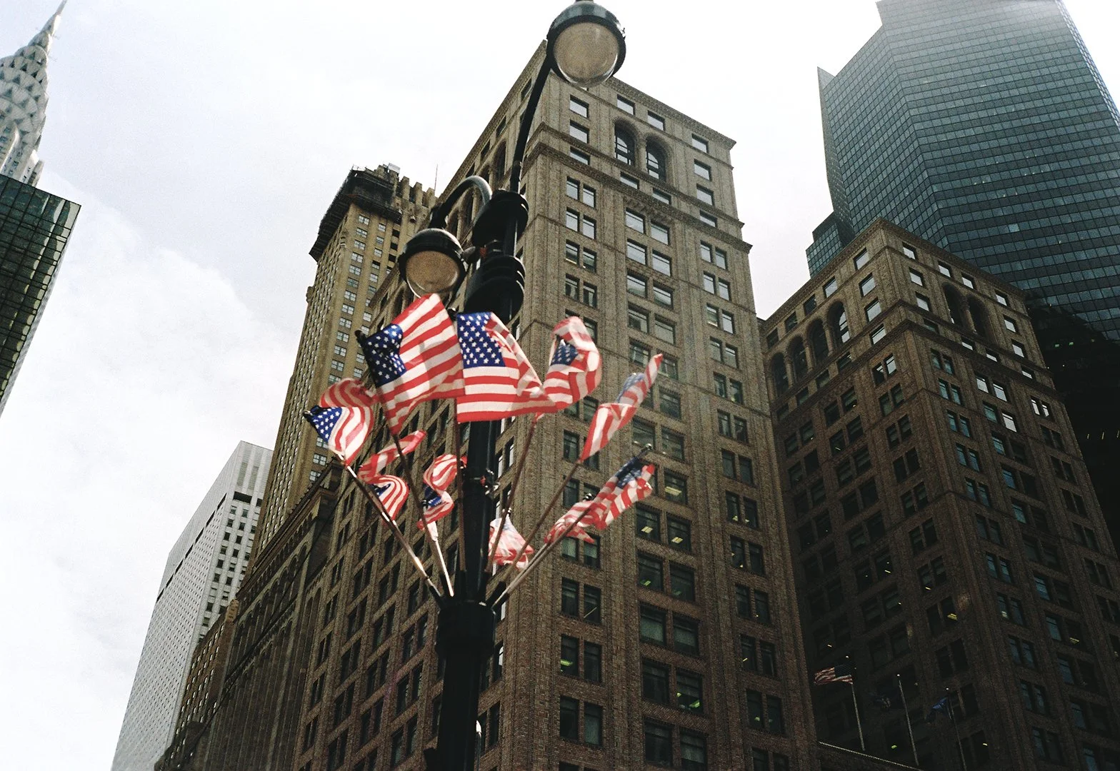A city street view with a pole decorated with multiple American flags, surrounded by tall skyscrapers and overcast sky.