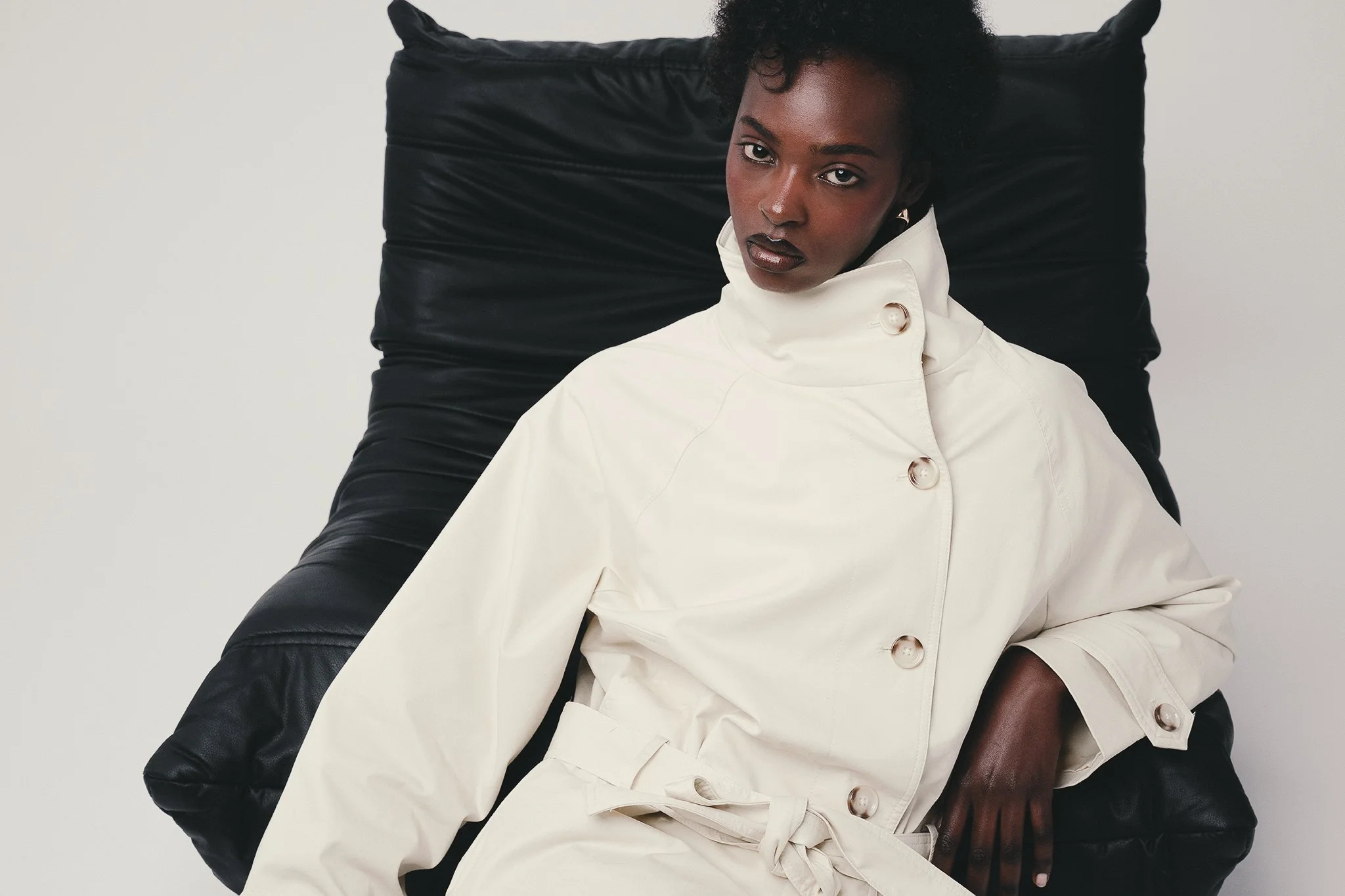 Portrait of a woman with dark skin, short curly hair, dark lipstick, wearing a white coat with large buttons, sitting on a black leather cushion or chair against a plain light background.